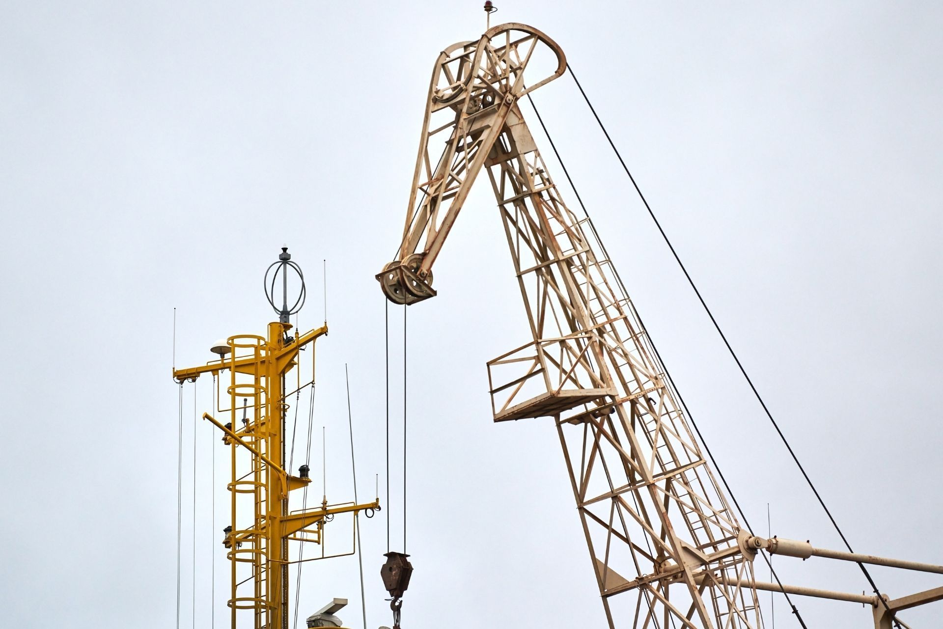 A large, tan industrial crane sits next to a tall, bright yellow mast against a pale gray sky.
