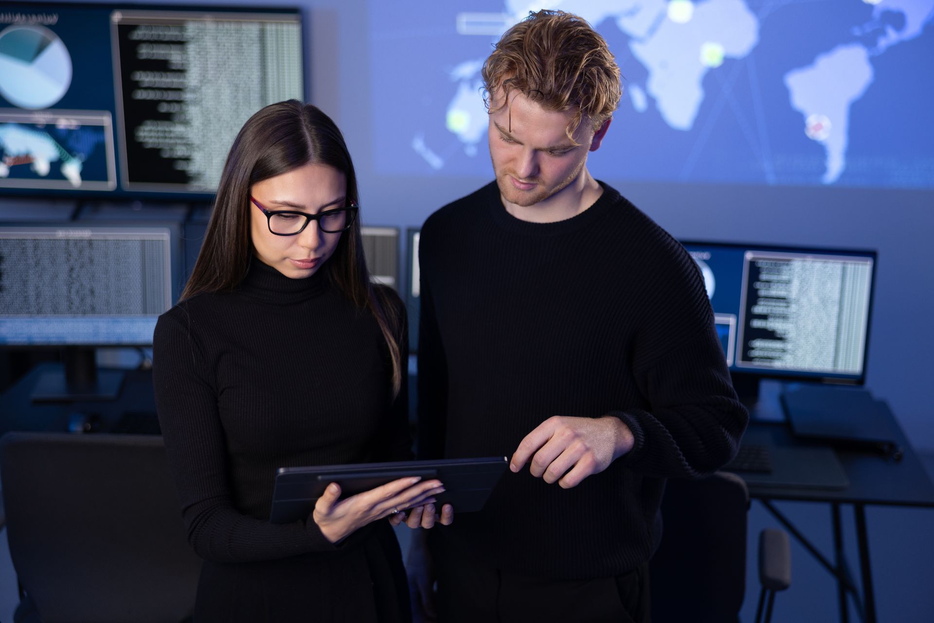 Two coworkers in black clothing reviewing a tablet in a dim tech office with monitors in the background