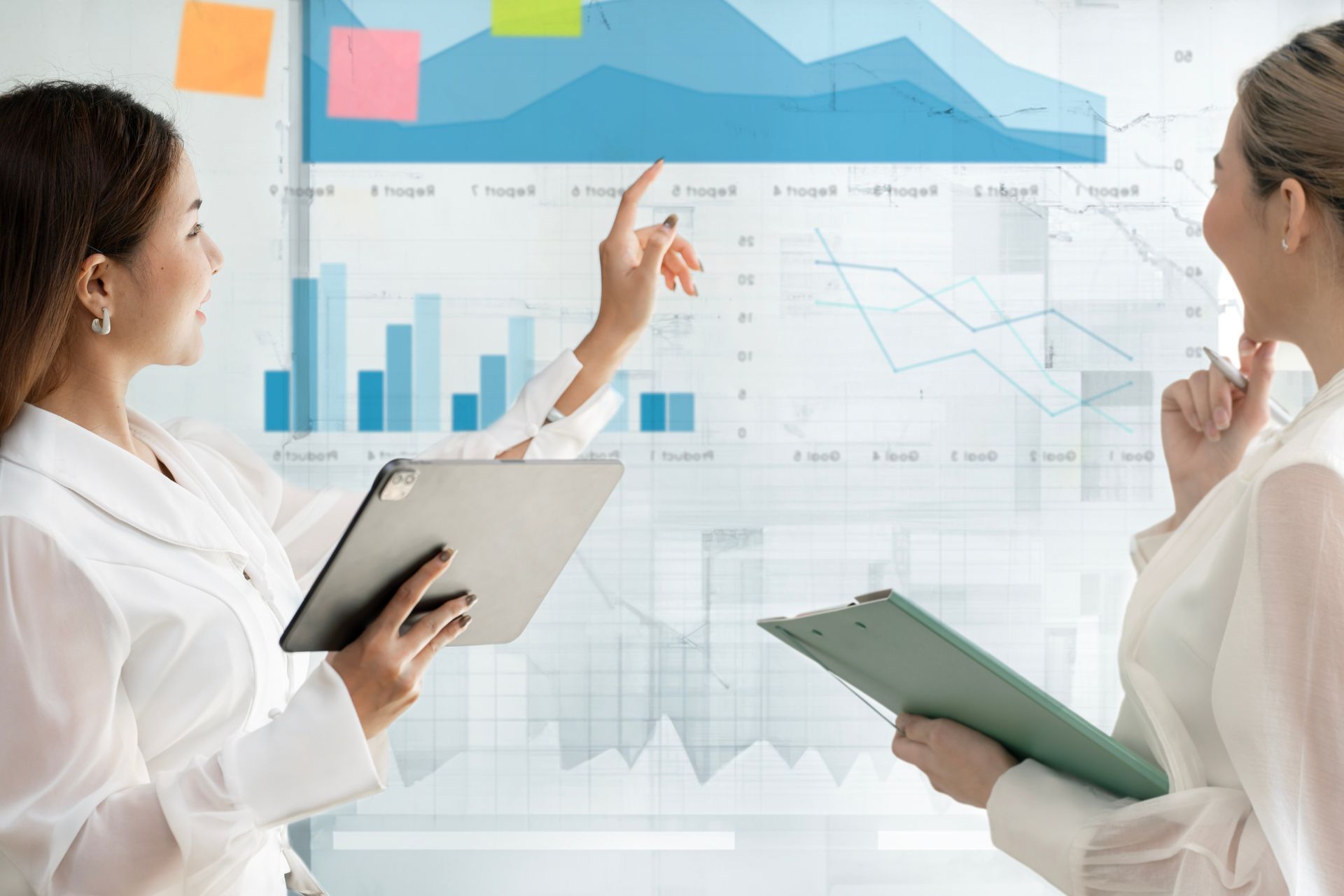 Two people in a bright office reviewing charts on a large glass screen with tablets.
