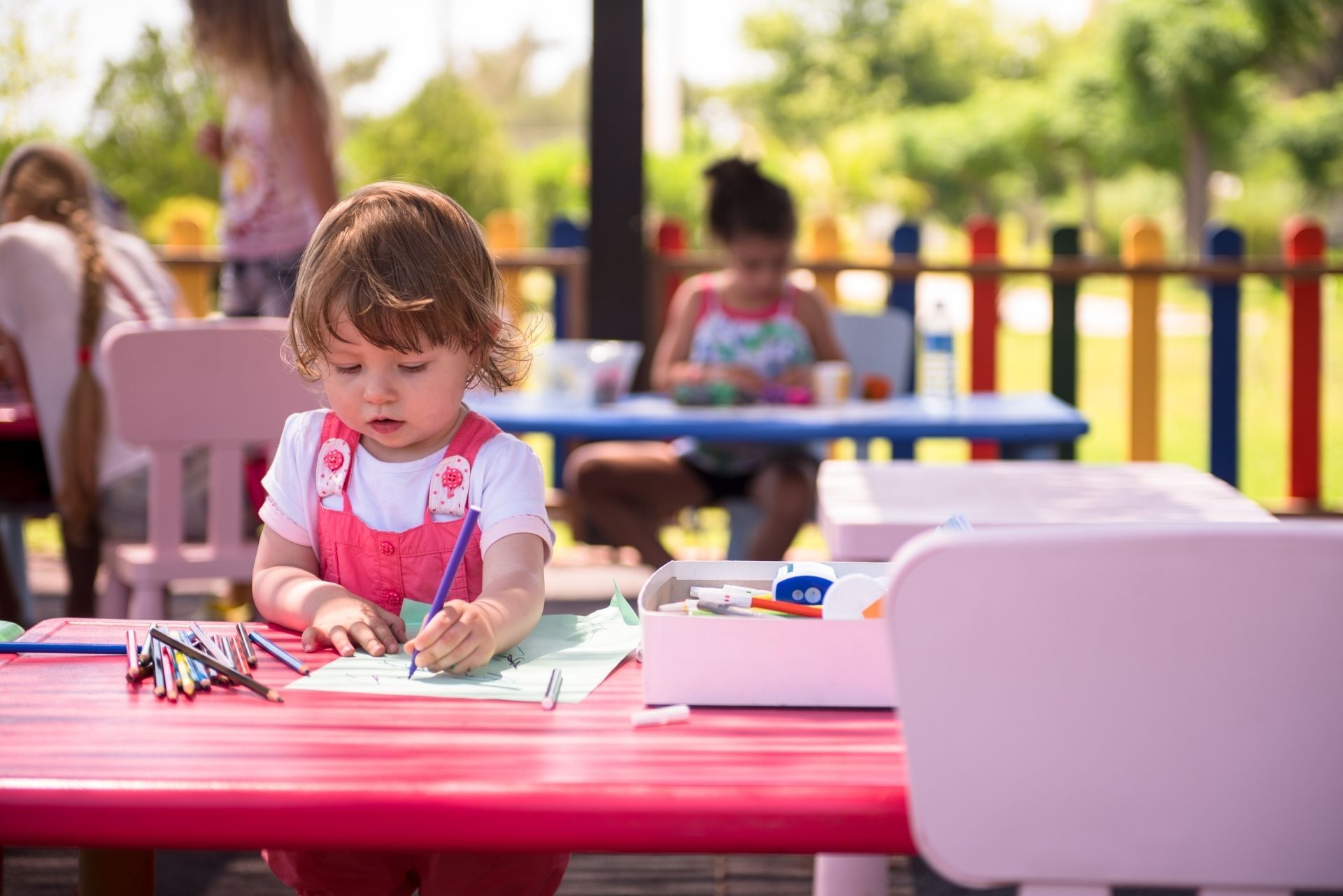 Child drawing at a pink table in a sunny outdoor play area with other children in the background