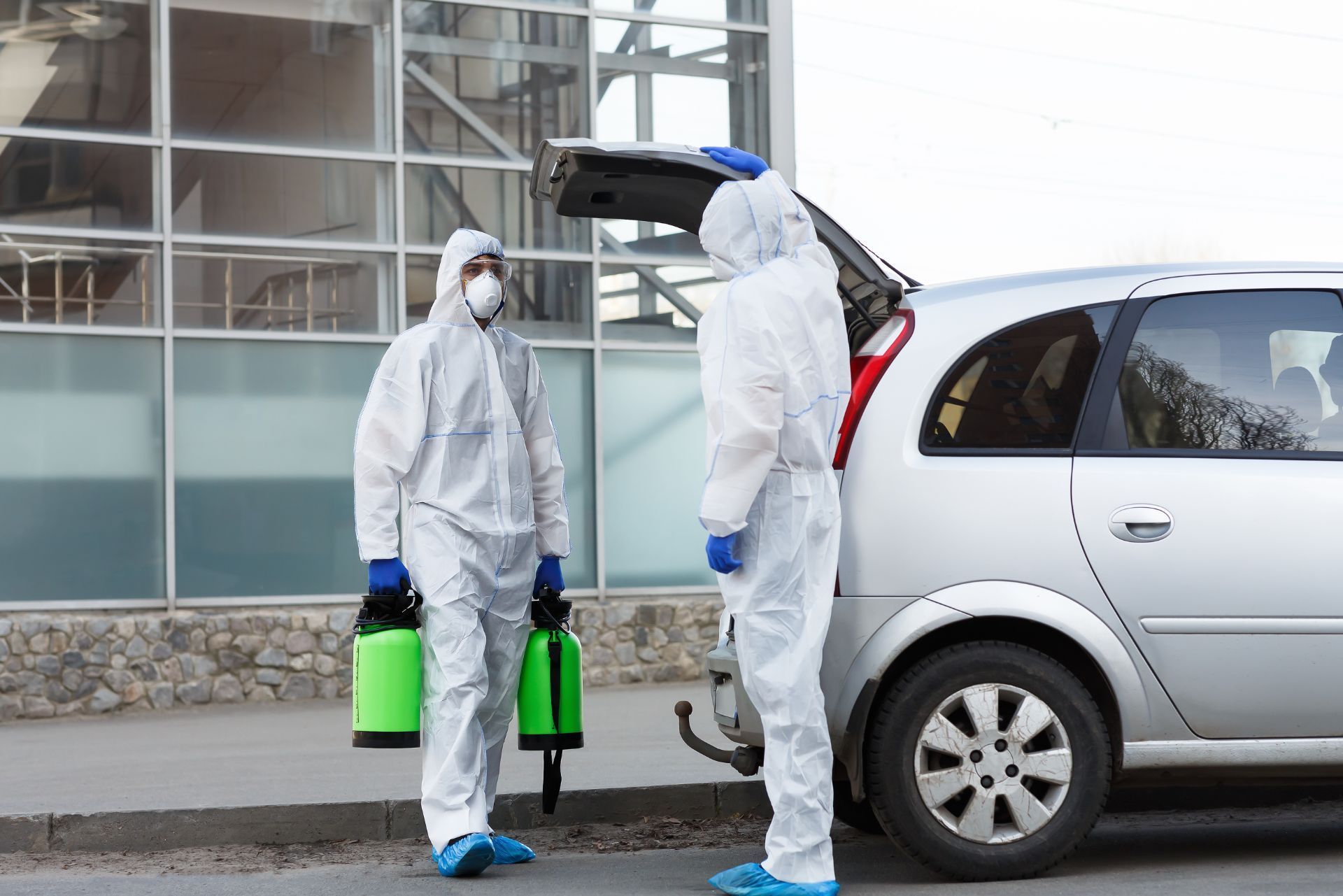 Two people in hazmat suits disinfecting a silver car outdoors with green sprayers