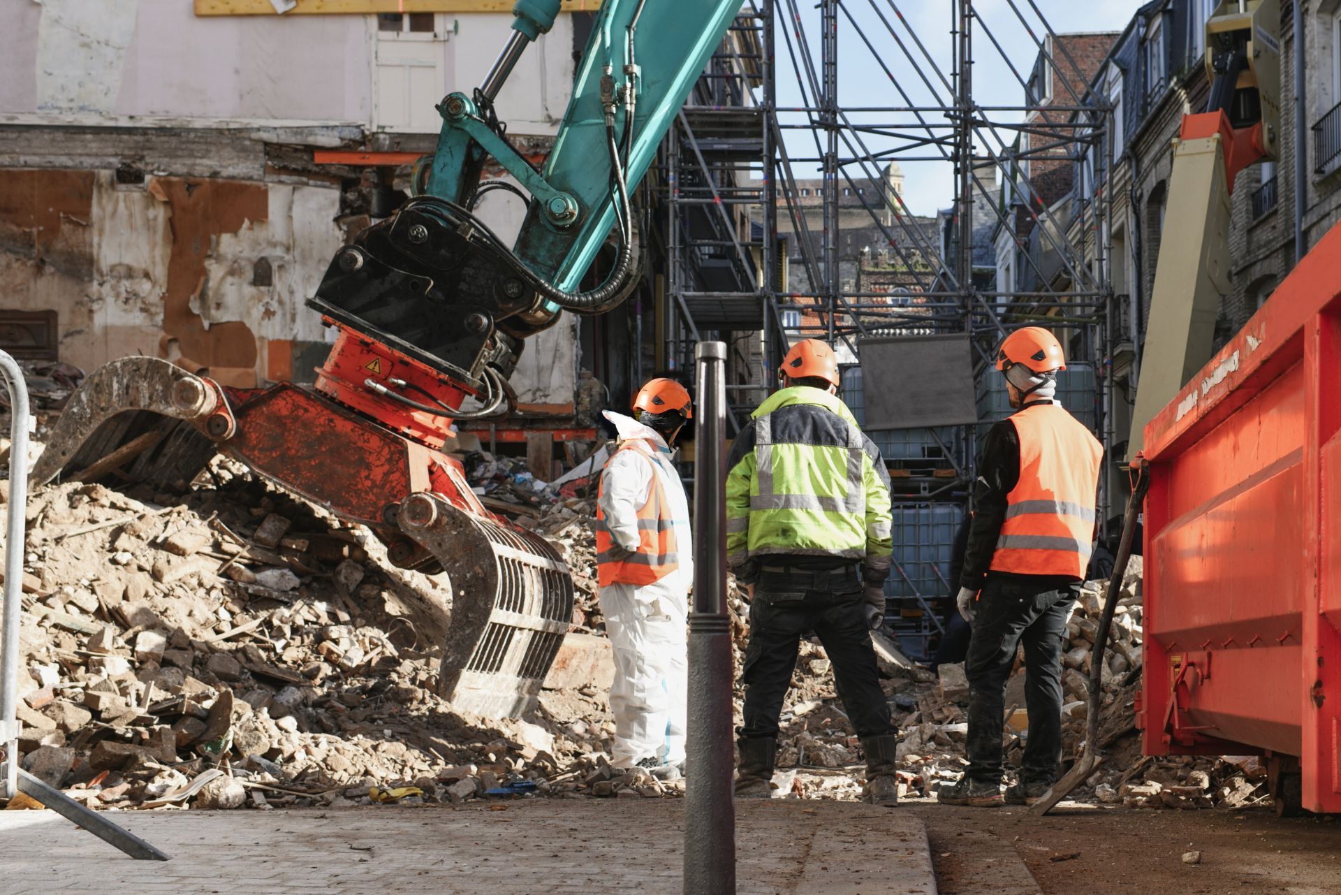 Construction workers in high-visibility gear observe a crane dismantling a building at a debris-filled site.