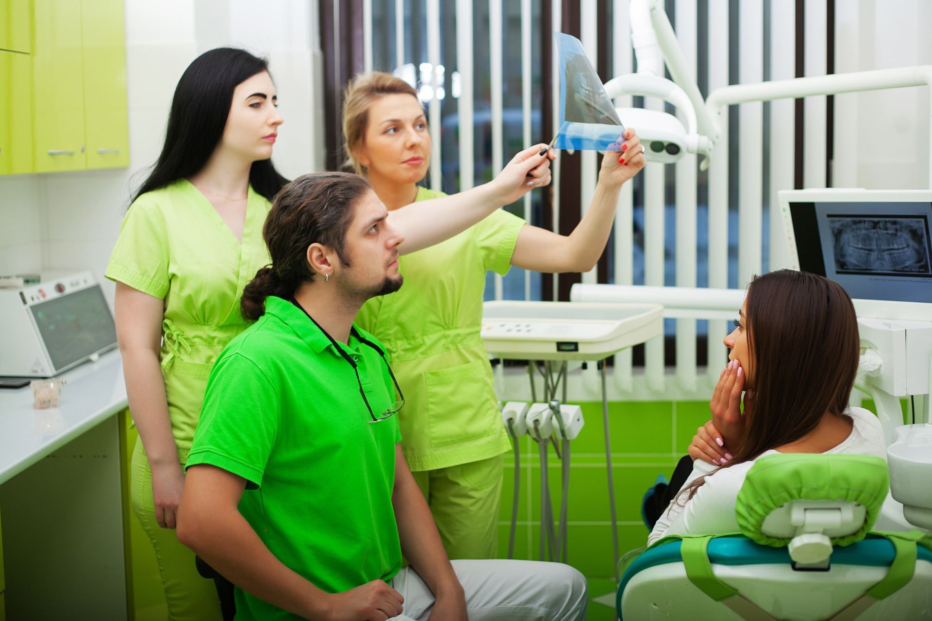 Dental team examining a patient in a green-and-white clinic, with one person adjusting equipment beside a monitor.