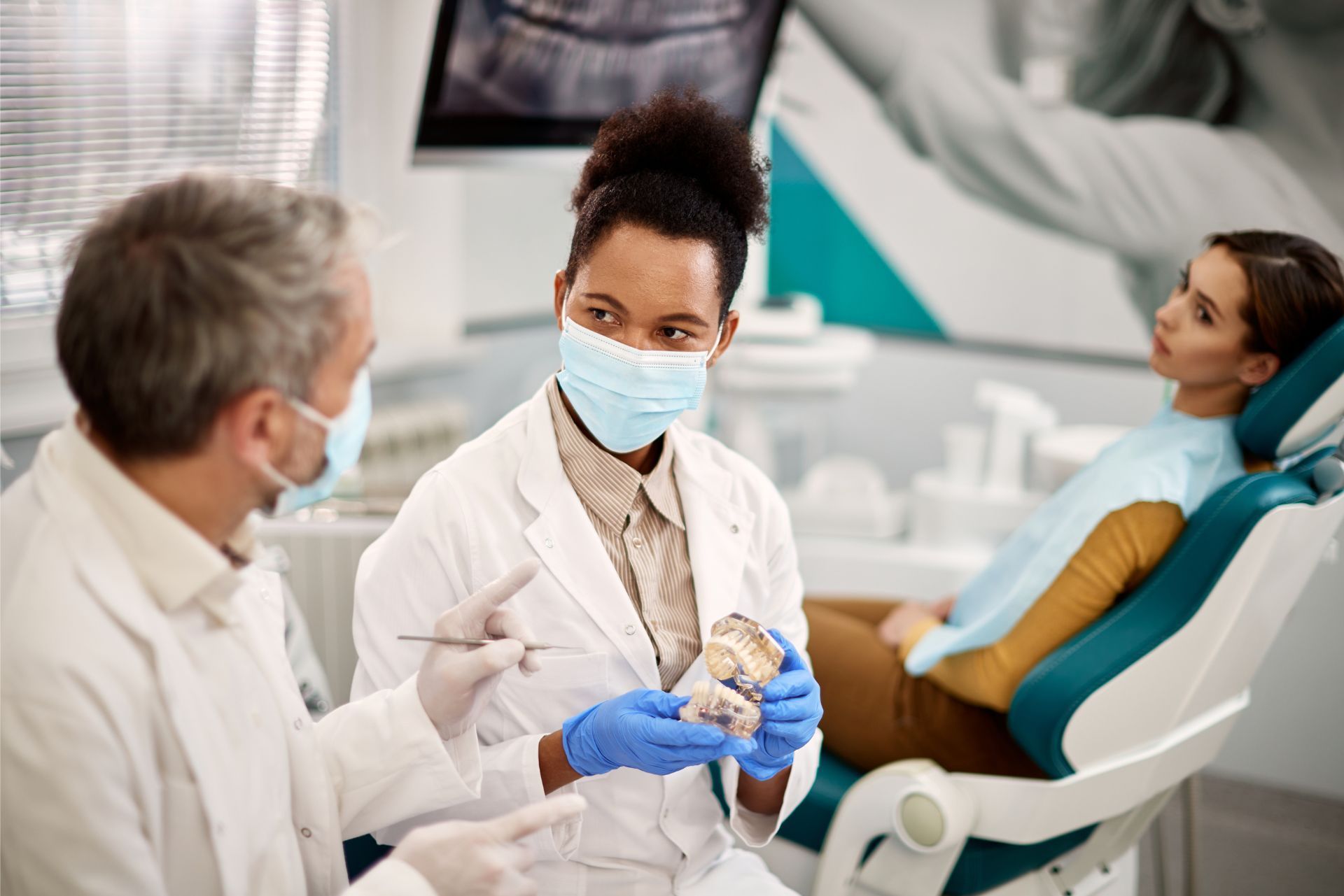 Dentist preparing a patient in a clinic while speaking with a seated colleague and a reclining patient in the background
