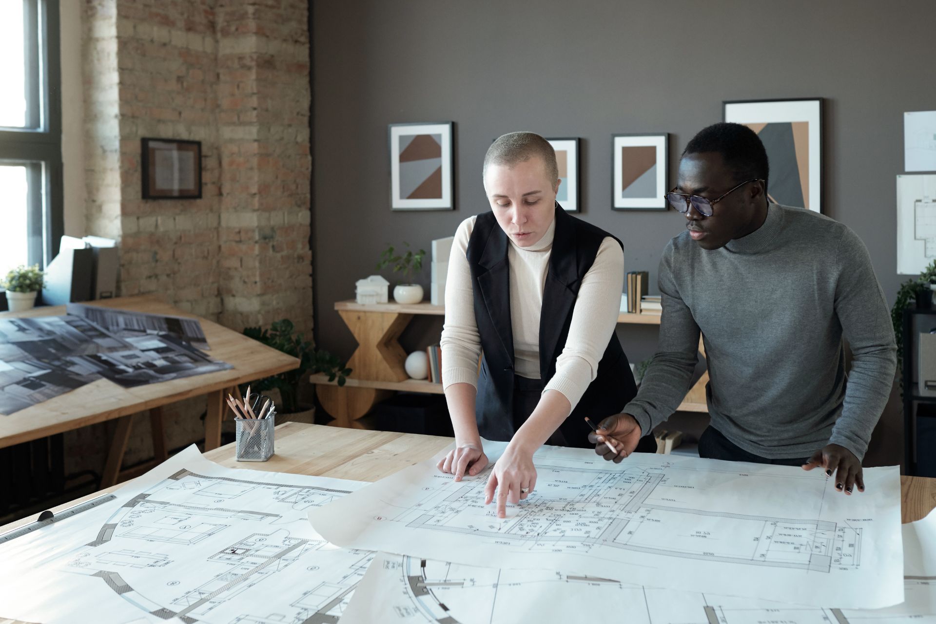 Two coworkers review large architectural blueprints on a table in a bright office.