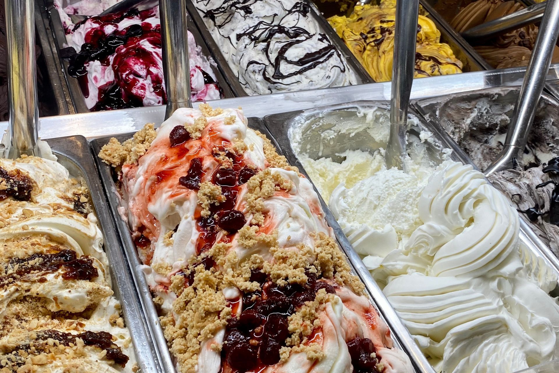 Assorted gelato flavors in a shop display case, including white swirls and berry and nut toppings.