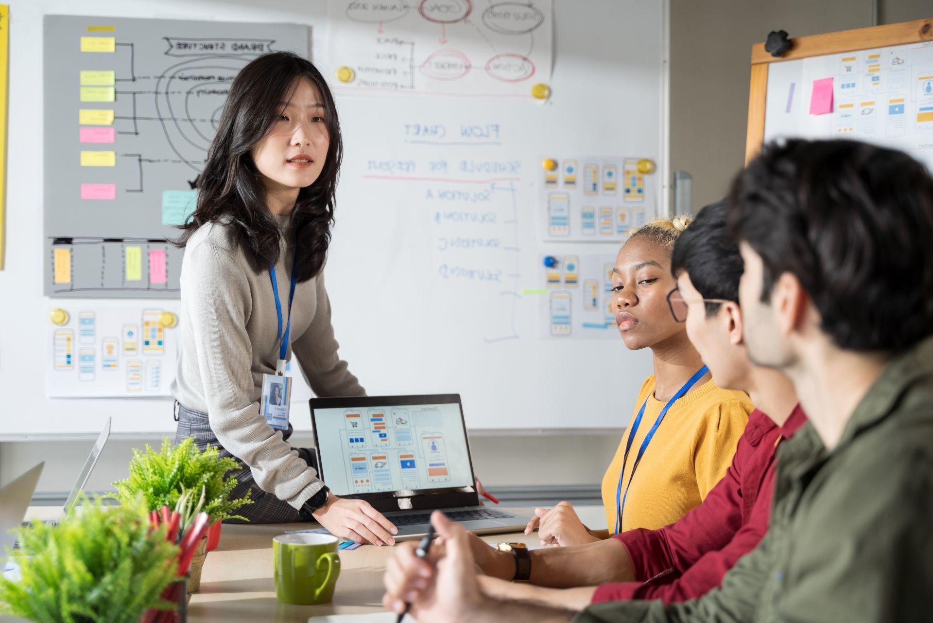 Team meeting around a laptop in a bright office, with a presenter standing by a whiteboard.