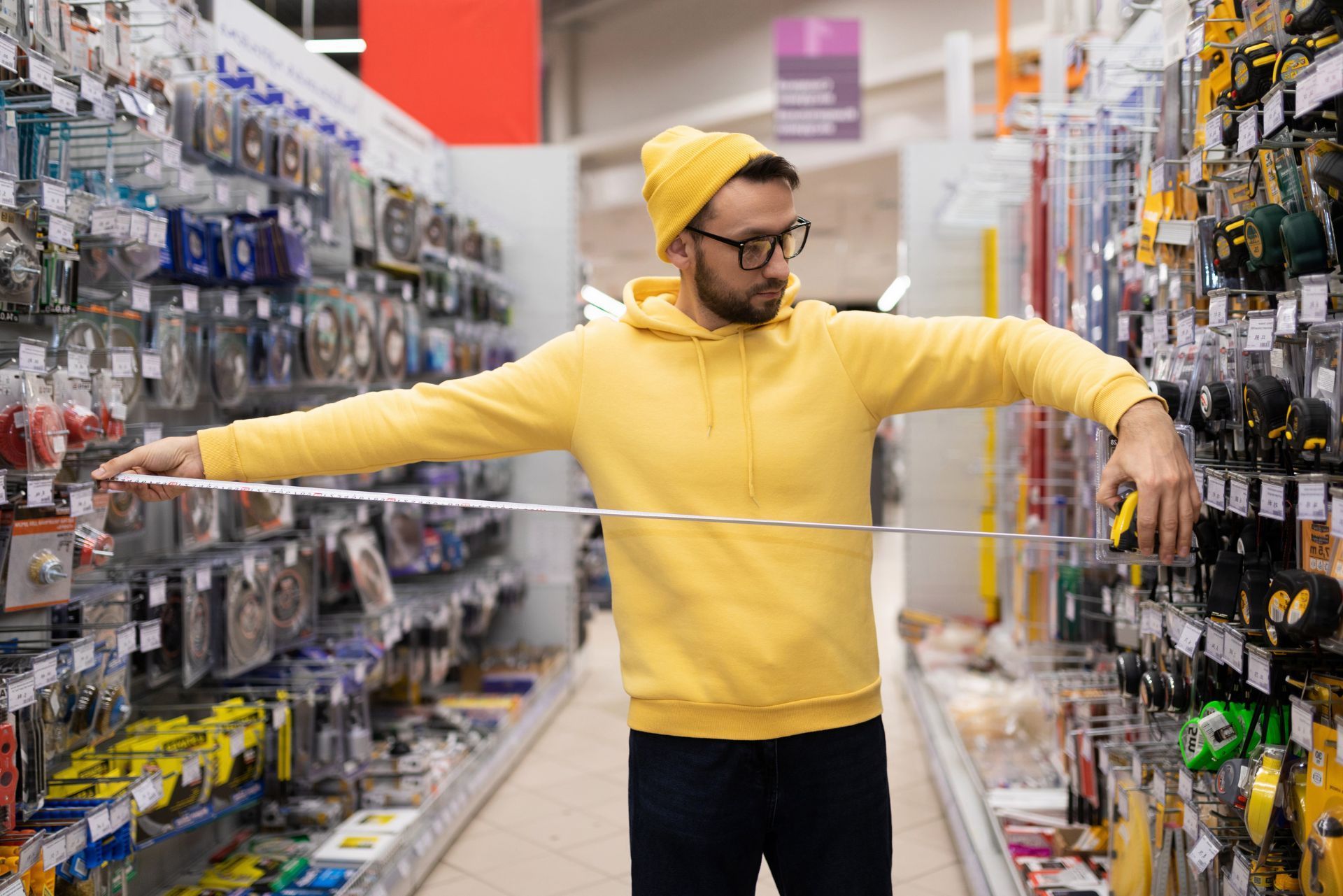 Man in a yellow hoodie holding a long ruler in a hardware store aisle