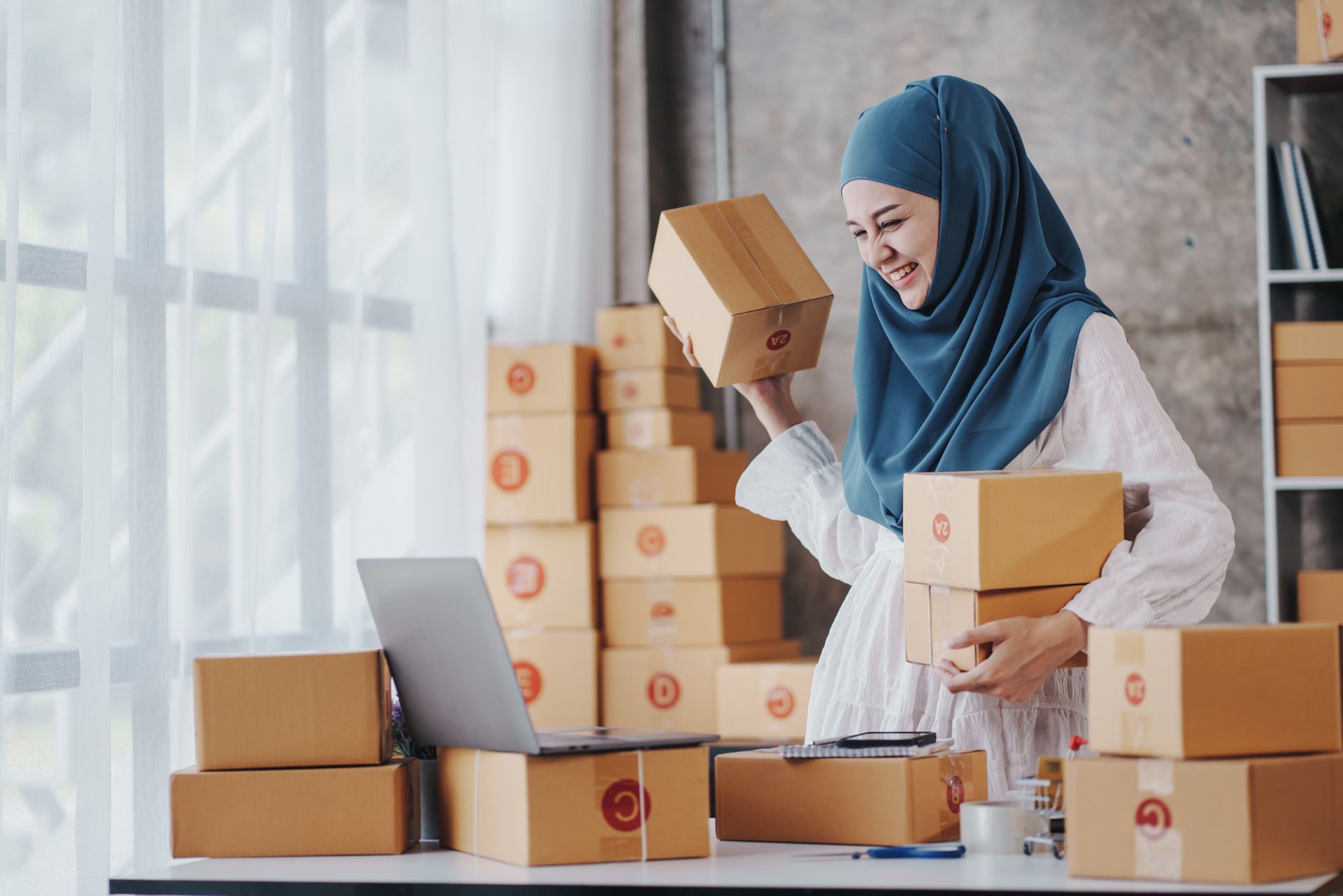 Woman packing cardboard boxes beside a laptop in a small home office