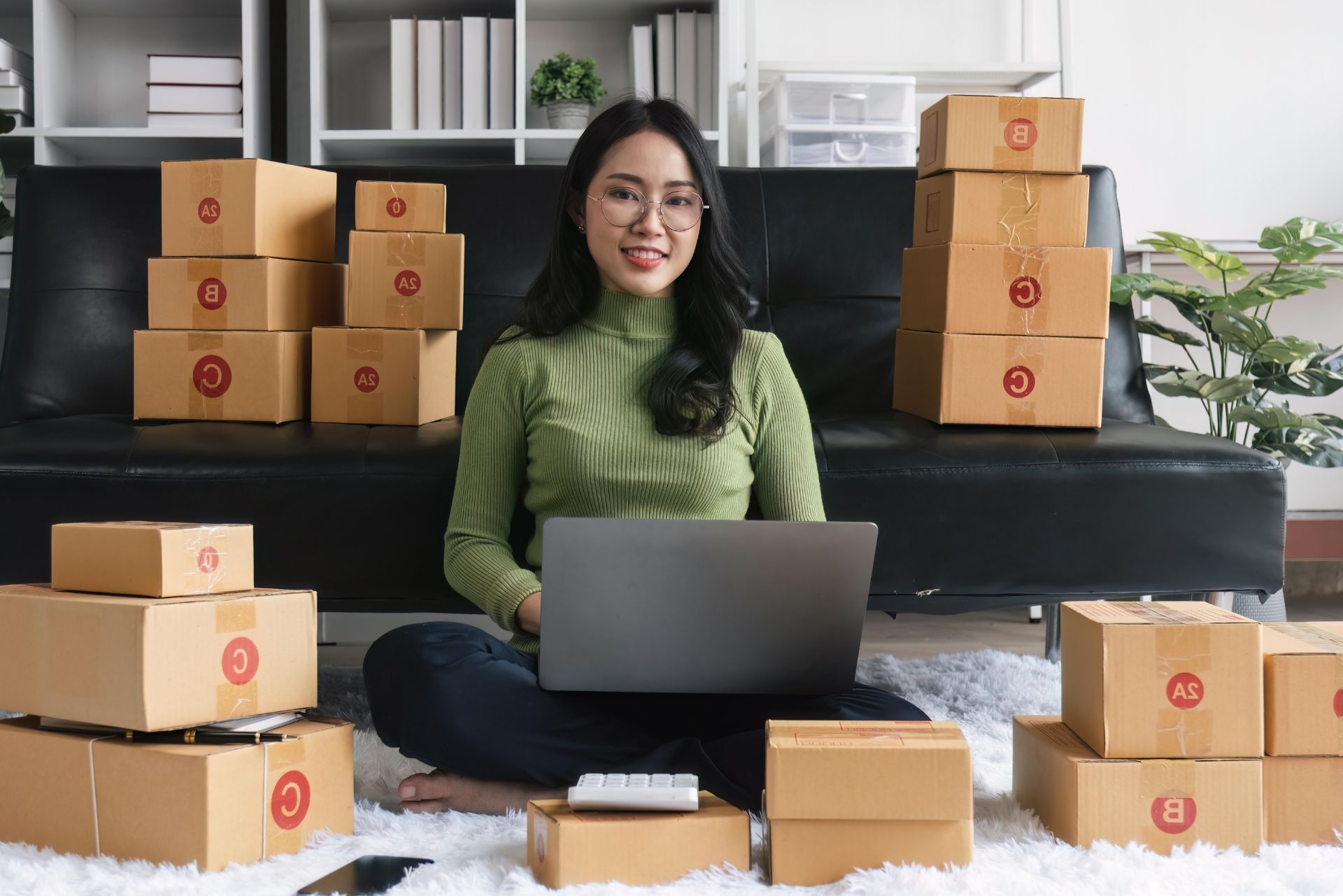 Person sitting with a laptop among stacked cardboard boxes in a home office or storage room