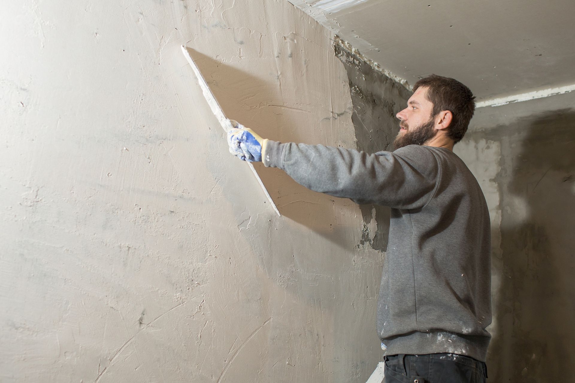 Man in a gray sweater plastering a wall with a trowel indoors