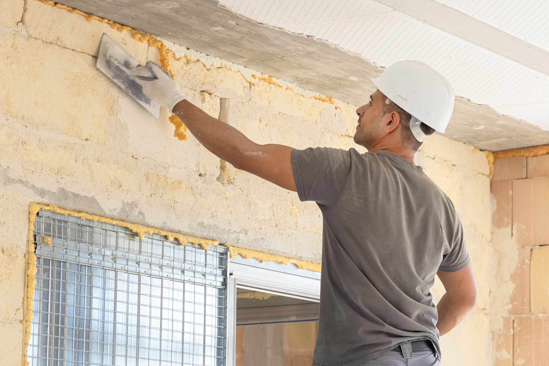 Worker in hard hat plastering a wall indoors with a trowel.