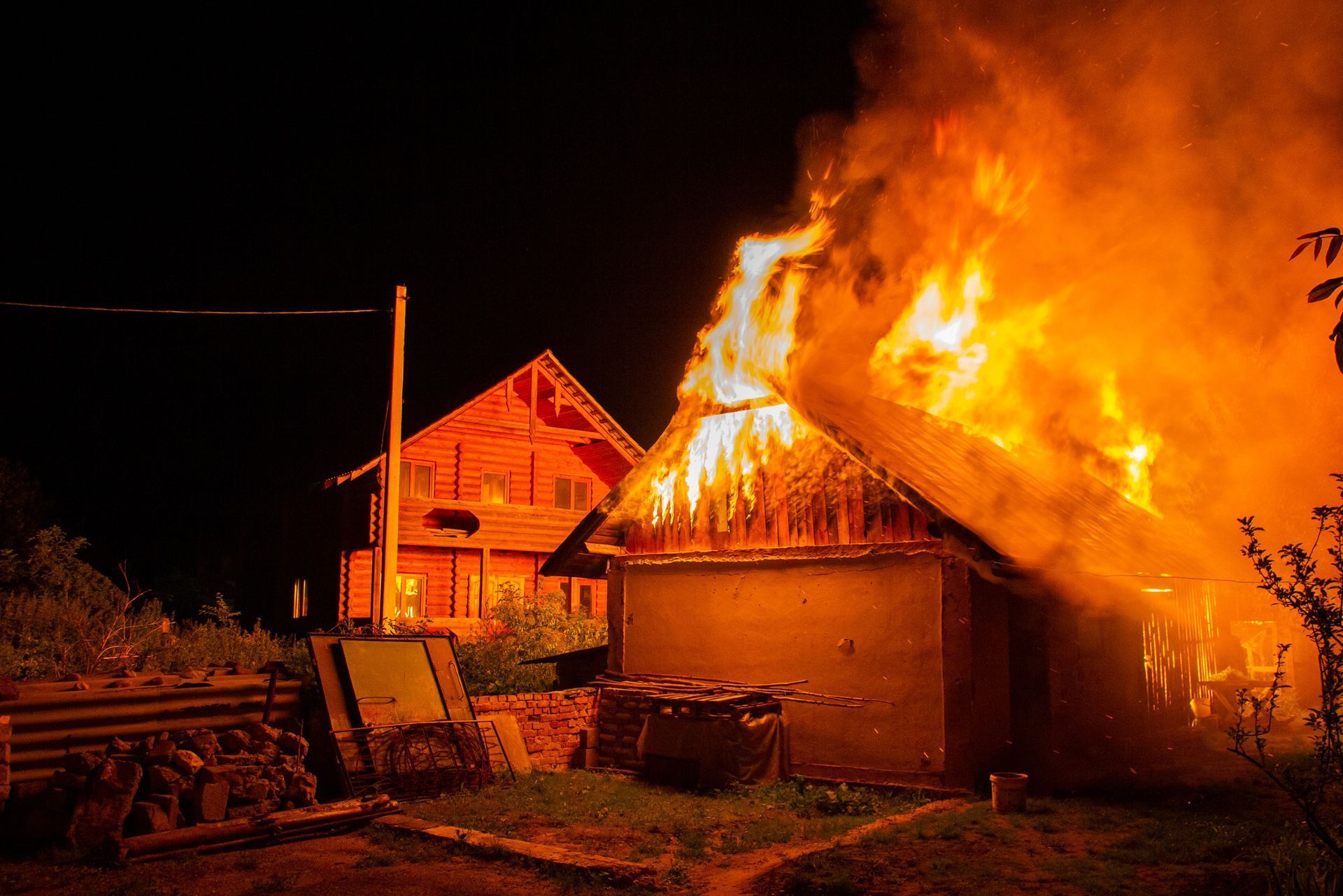 House roof engulfed in bright flames at night, with another house and yard equipment nearby