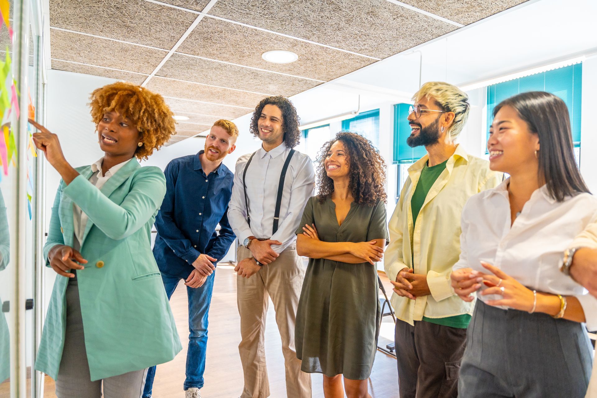 Five coworkers chatting in a bright office hallway, one holding a tablet and others smiling.