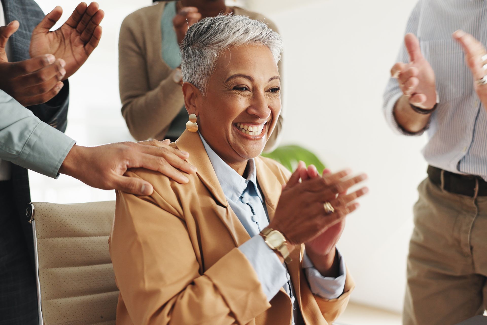 Smiling woman in a tan blazer claps while others applaud around her in a bright office setting
