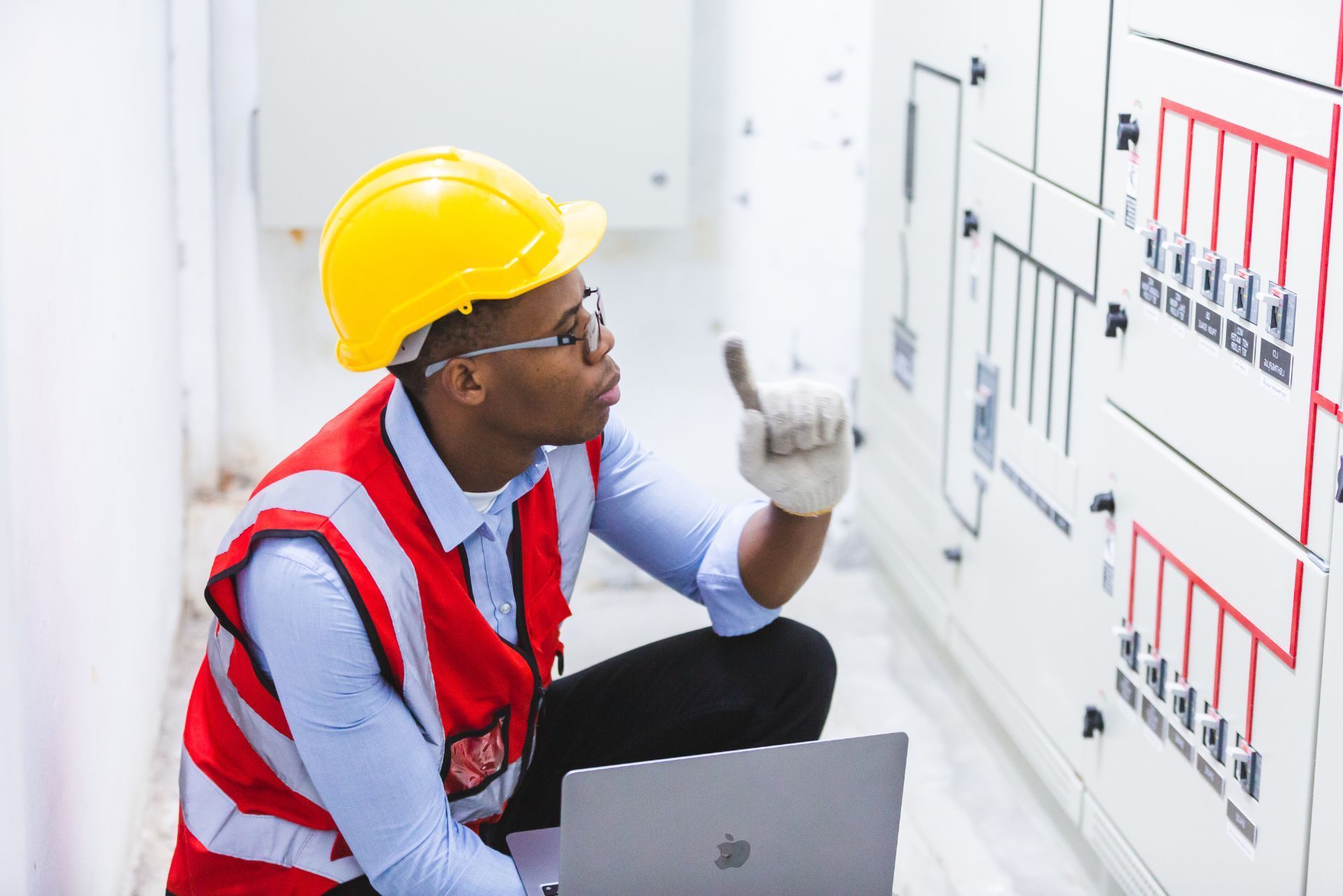 Technician in hard hat inspecting an electrical control panel with a laptop beside him