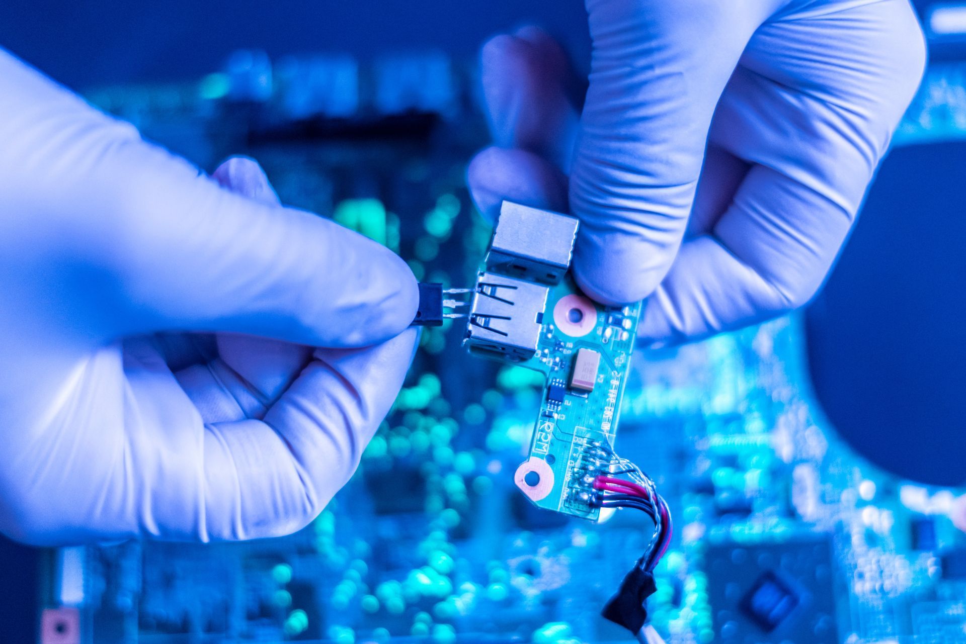 Gloved hands soldering a small circuit board under blue workshop lighting