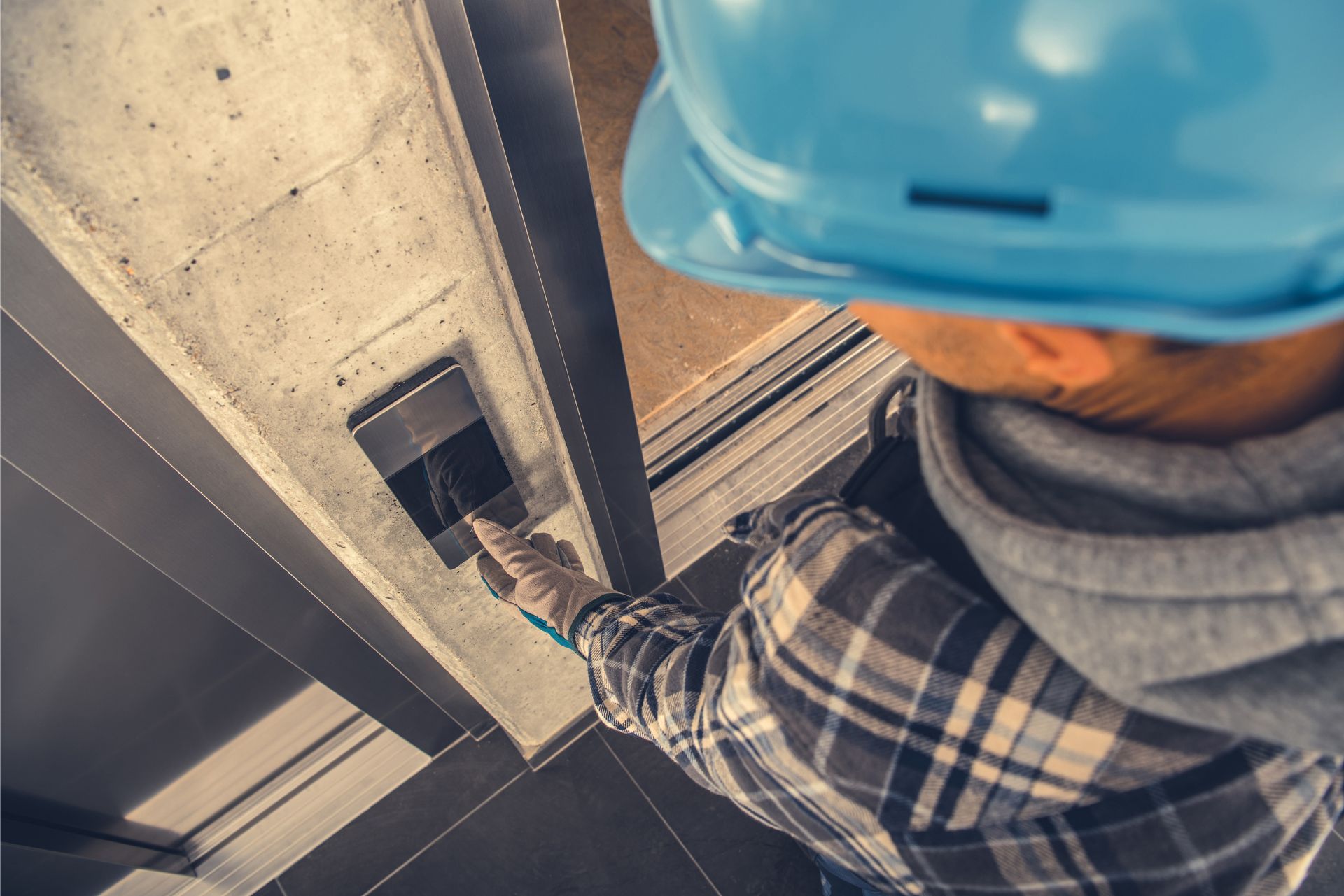 A worker in a blue hard hat and plaid shirt presses a key card against a door access reader in a modern building.