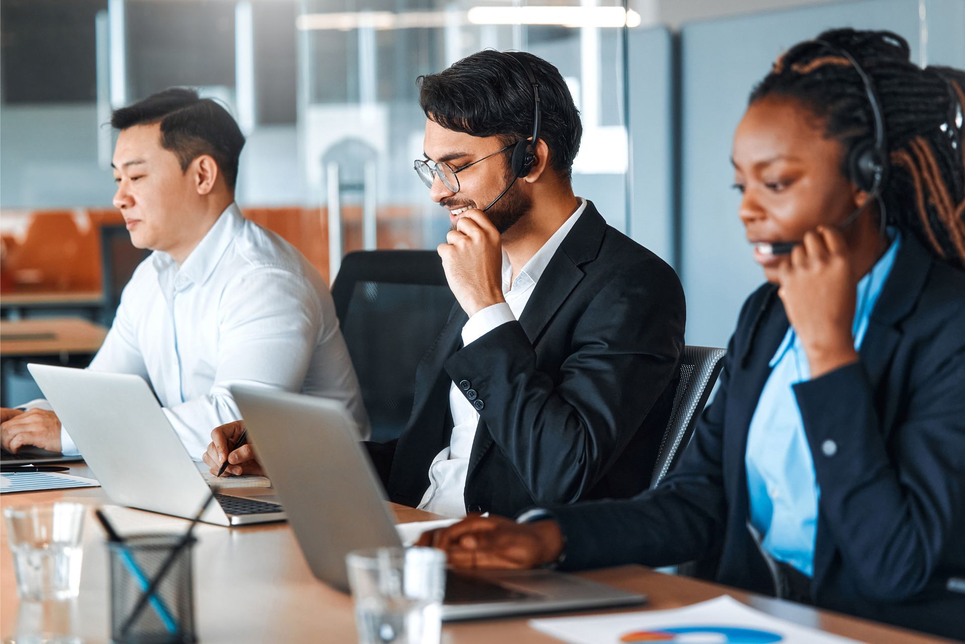 Three coworkers in business attire work on laptops during a meeting at a conference table.