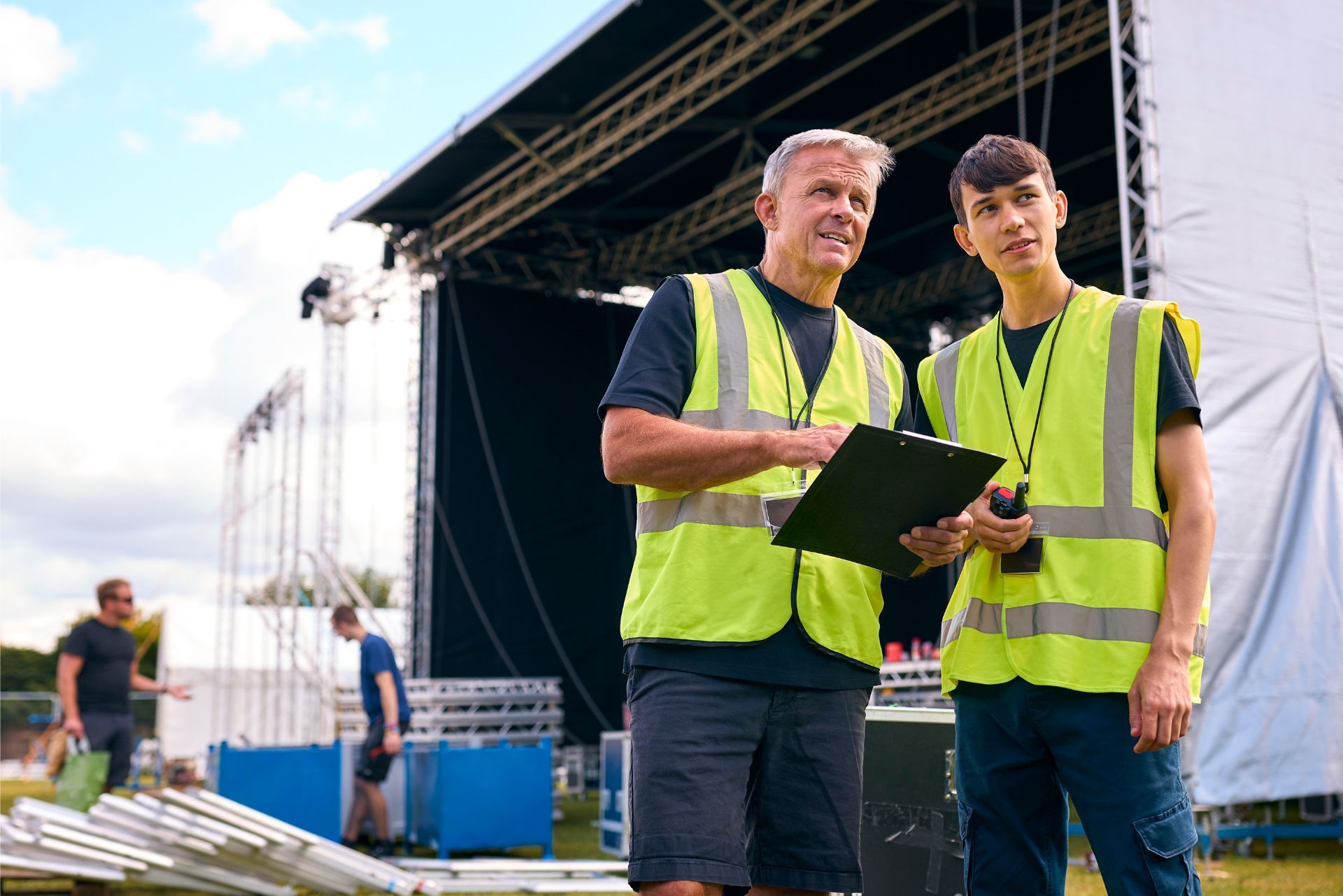 Two workers in yellow vests reviewing plans at an outdoor stage setup