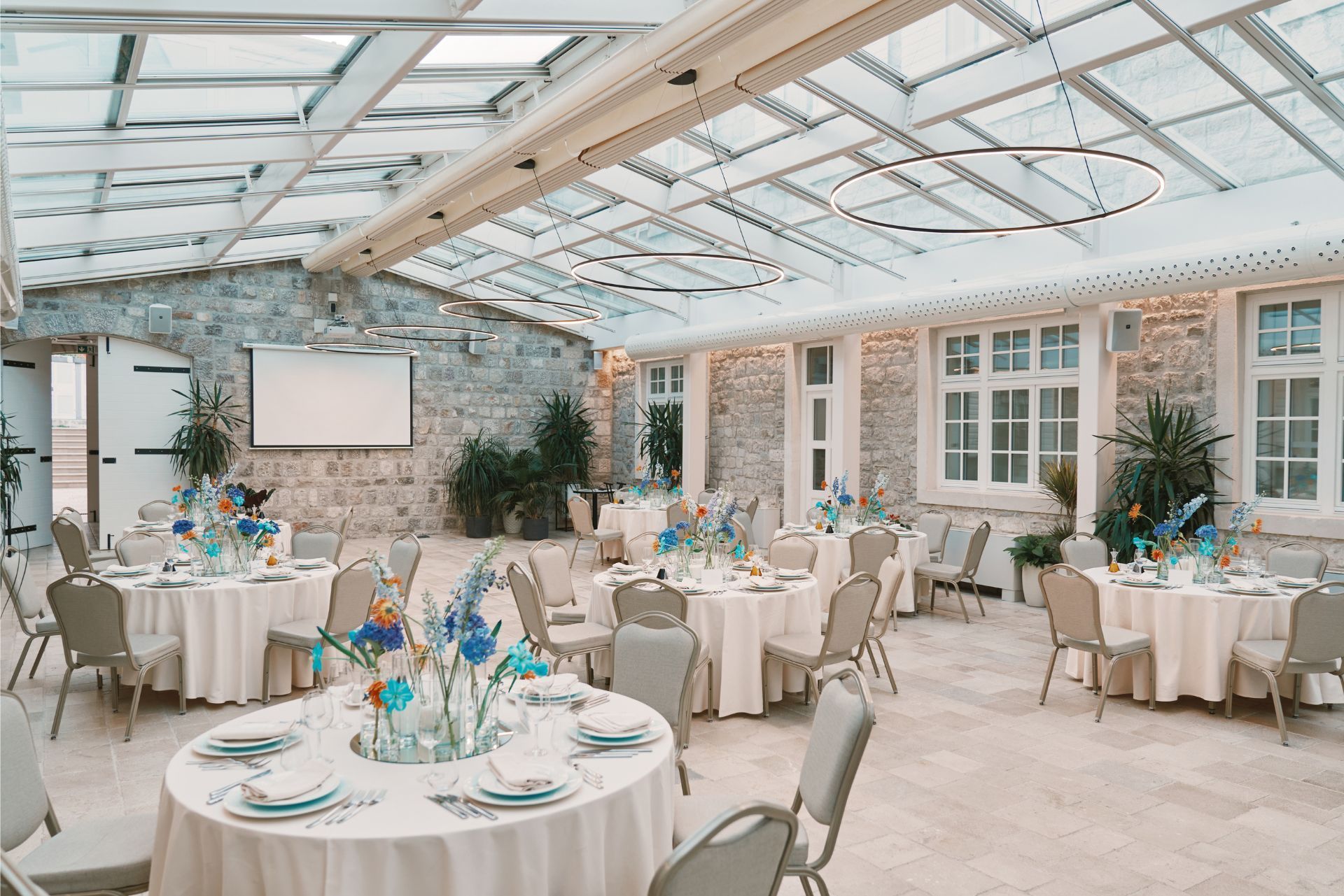 An indoor event space with round tables, white tablecloths, chairs, stone walls, and a glass ceiling.