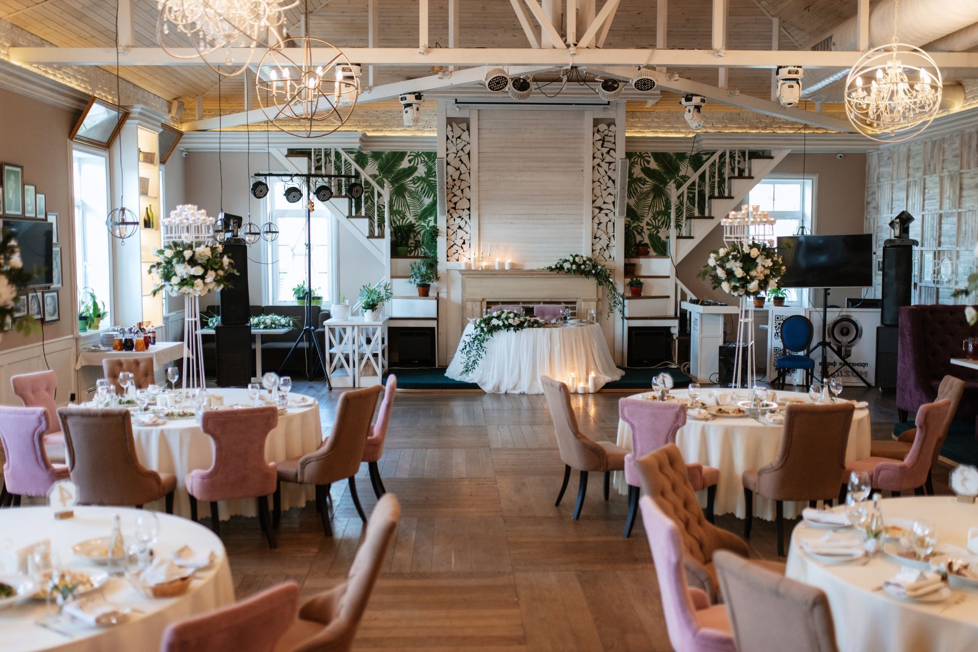 A banquet hall prepared for a wedding with round tables, floral centerpieces, and a draped sweetheart table at the back.