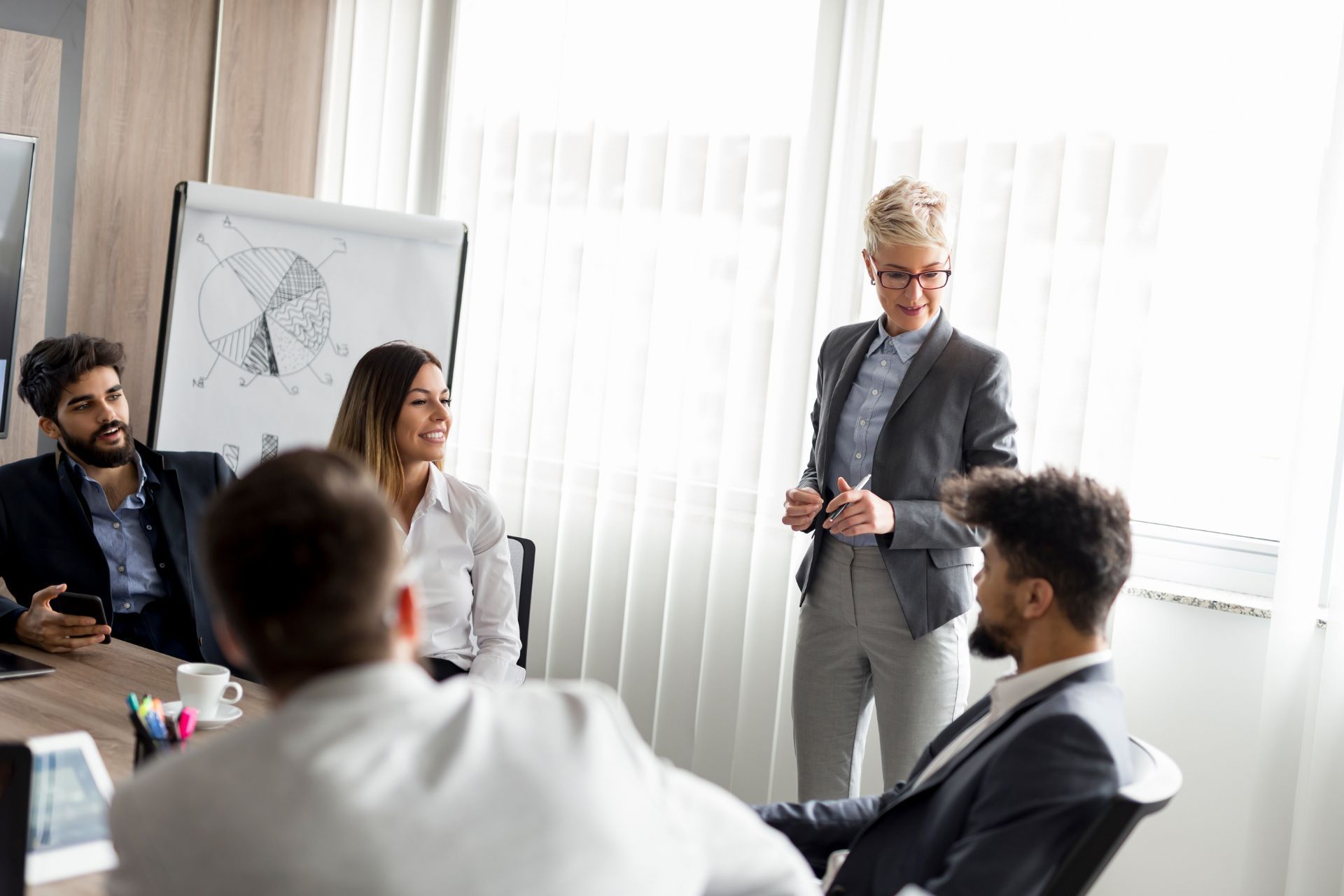 Business meeting in a bright conference room, with a presenter standing by a flip chart and colleagues seated nearby.