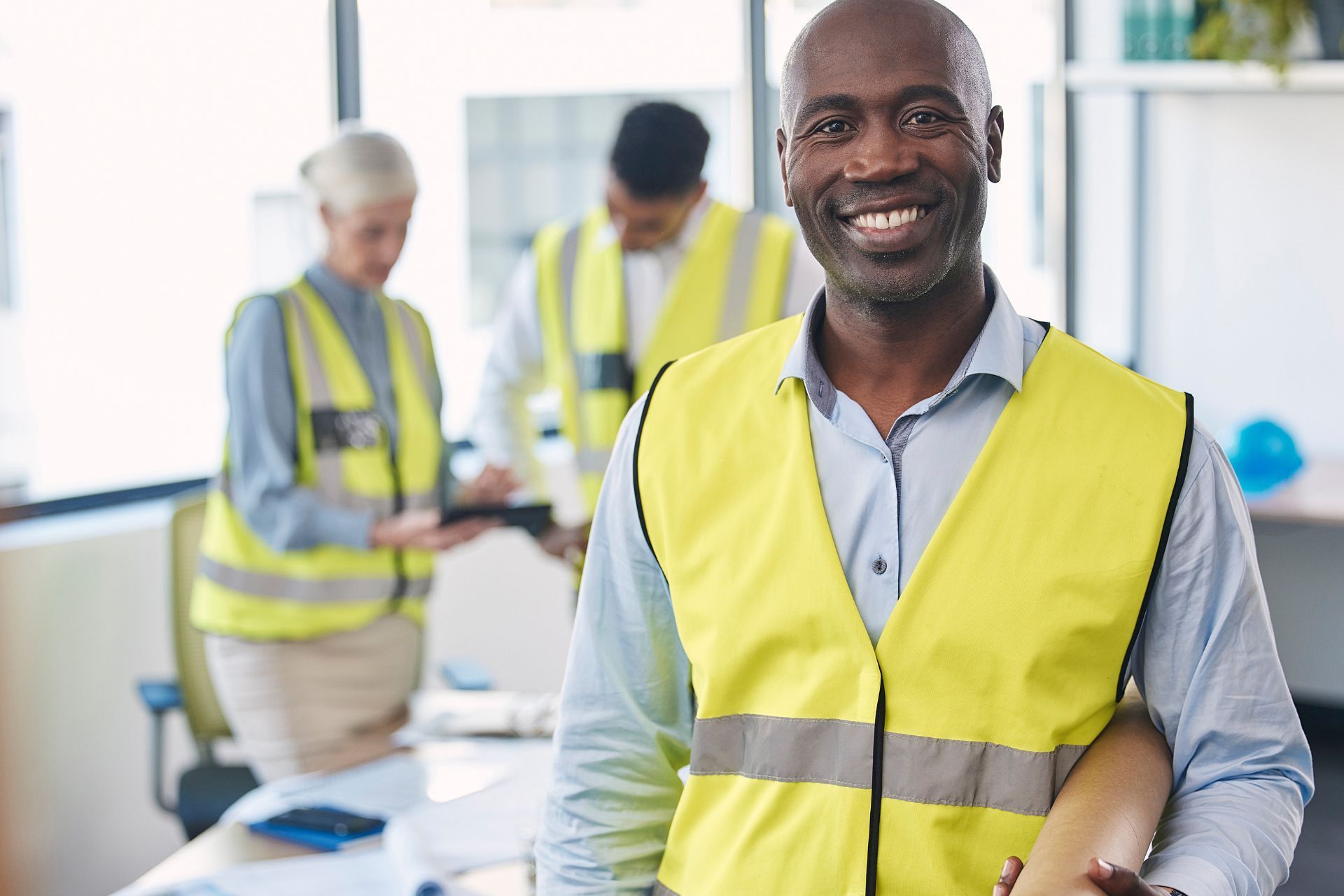 Smiling worker in yellow safety vest in an office, with two colleagues in the background