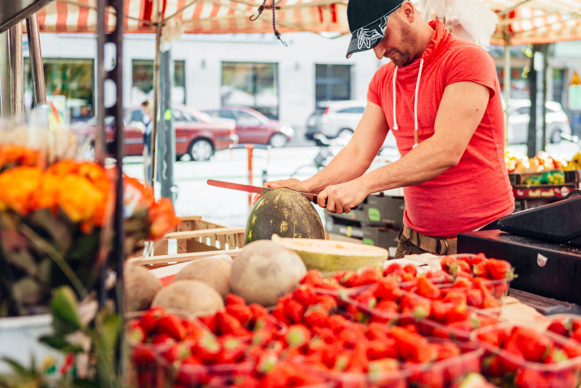 Vendor slicing a watermelon at an outdoor market, with strawberries and melons in the foreground.