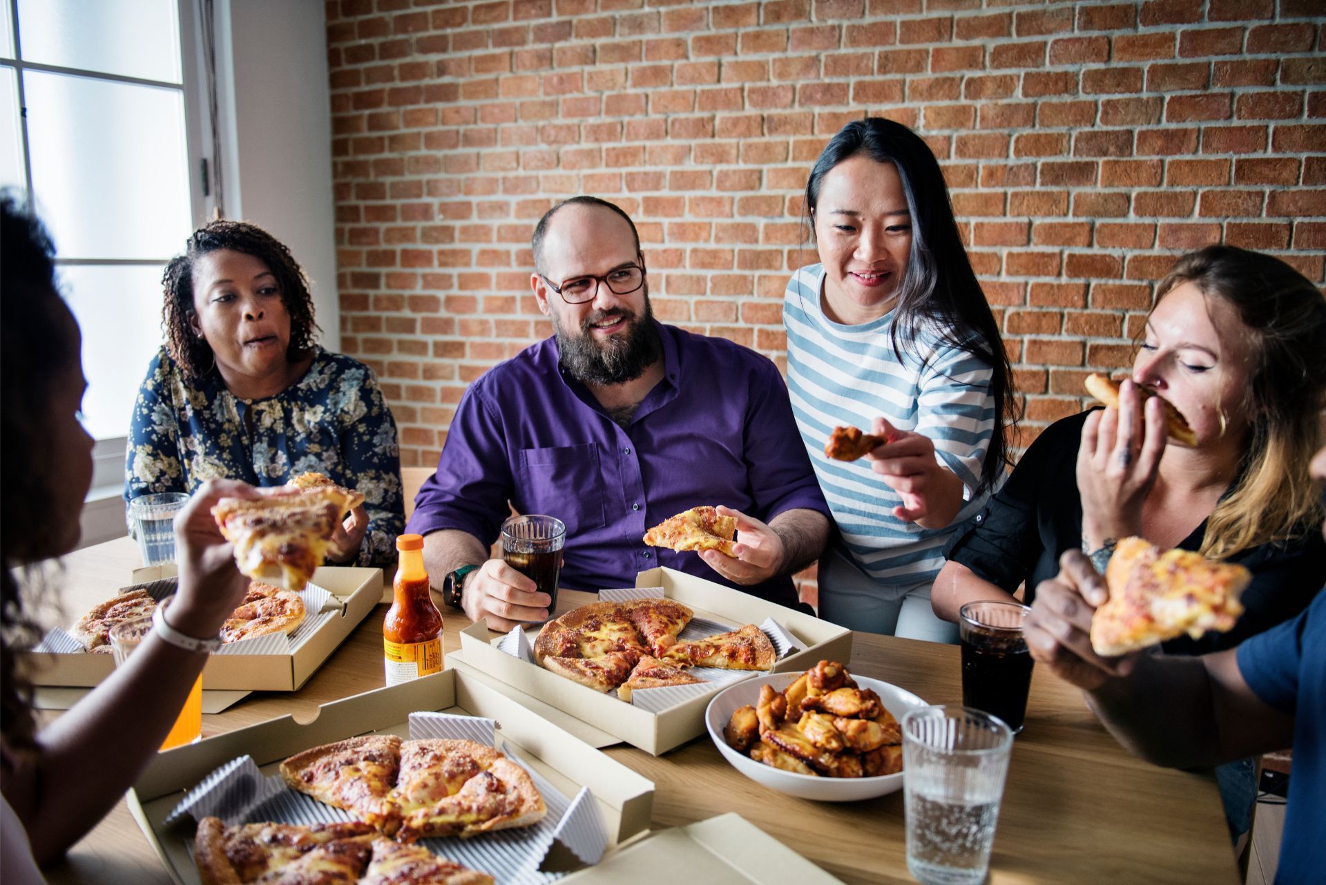 Five people eating pizza and chicken wings together at a wooden table in a room with a brick wall.