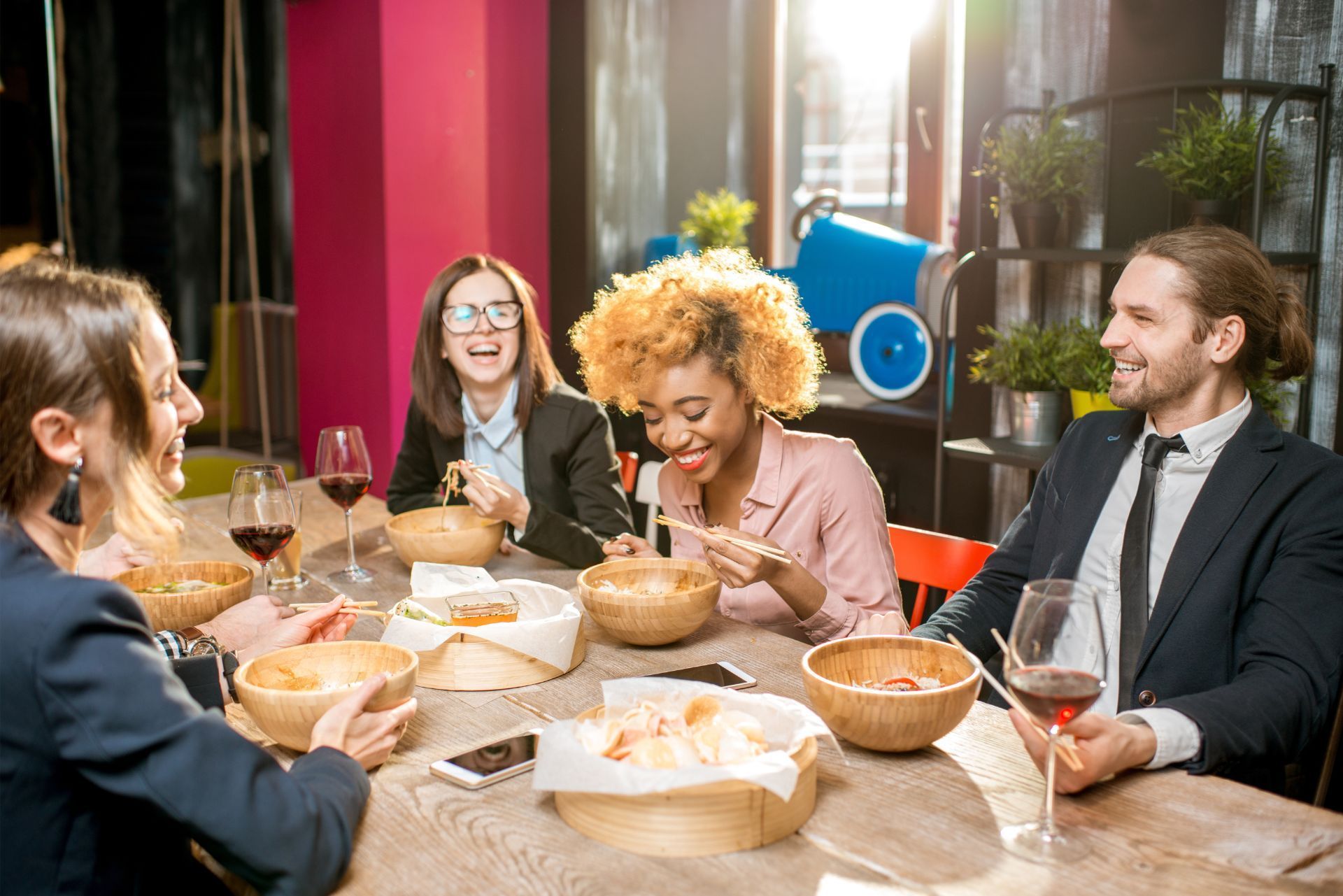 Four colleagues smile and eat together at a wooden table in a brightly lit, modern office space.