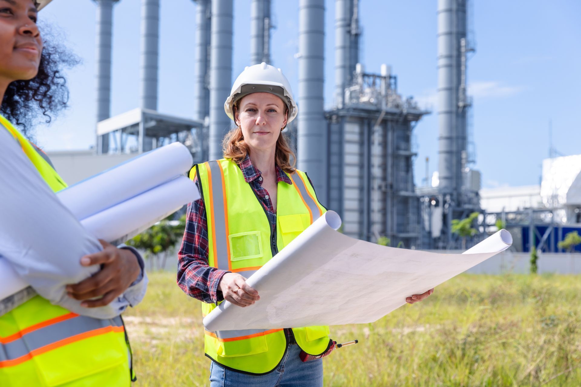 Two engineers in safety vests holding blueprints at an industrial refinery site