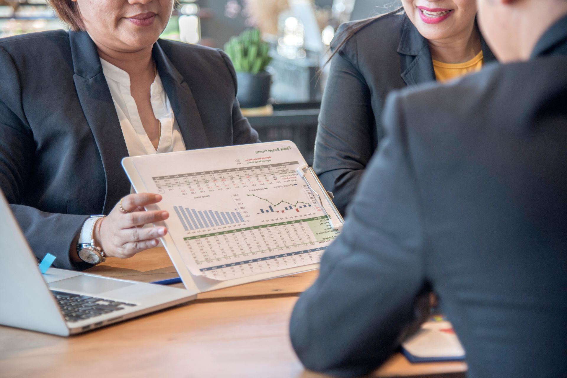 Three people in business attire review a printed financial report with charts and data at a table with a laptop.