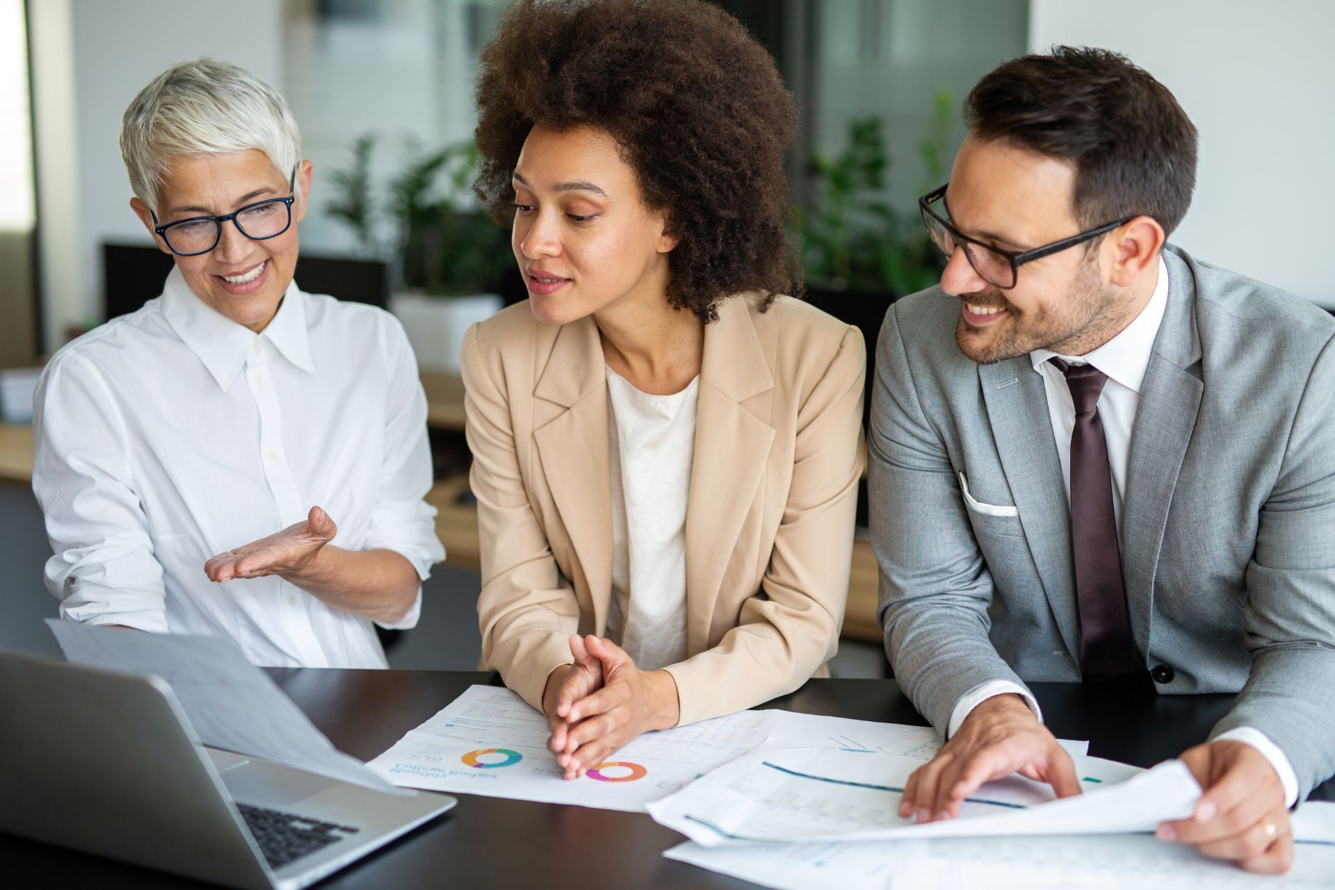 Three professionals in business attire collaborating over documents and a laptop at a table in an office setting.
