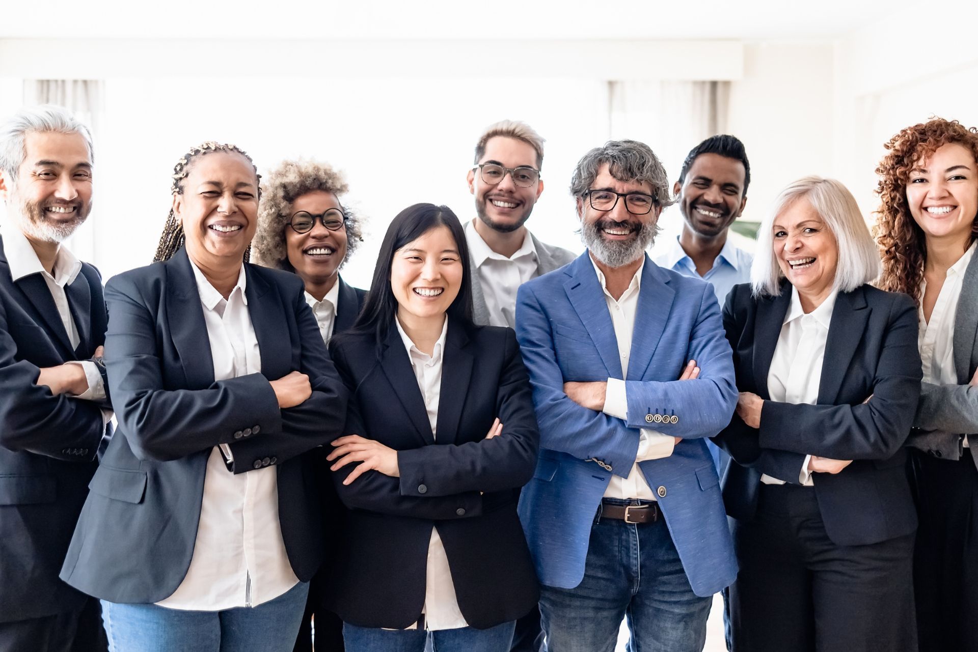 A diverse group of eight smiling professionals stand together in an office, most with arms crossed, wearing business attire.