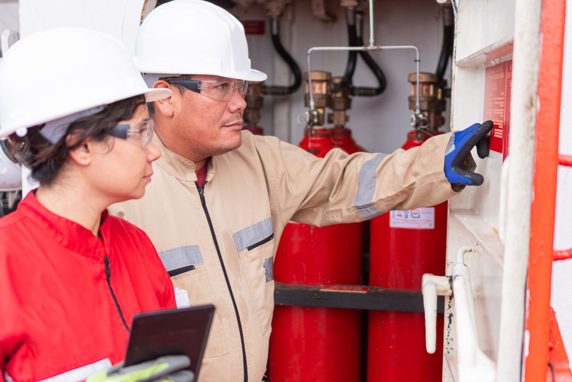 Two workers in hard hats inspect equipment in an industrial facility, one pointing at a red tank.