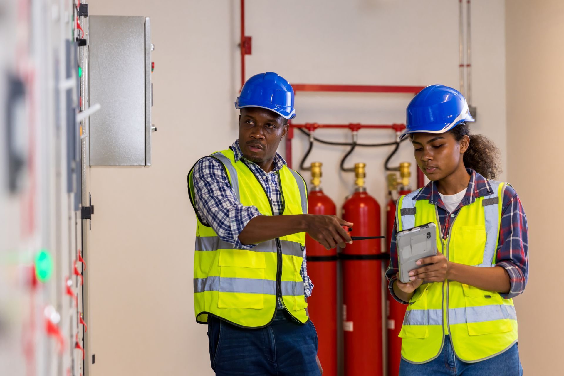 Two workers in blue hard hats and yellow safety vests inspecting fire extinguishers in a hallway
