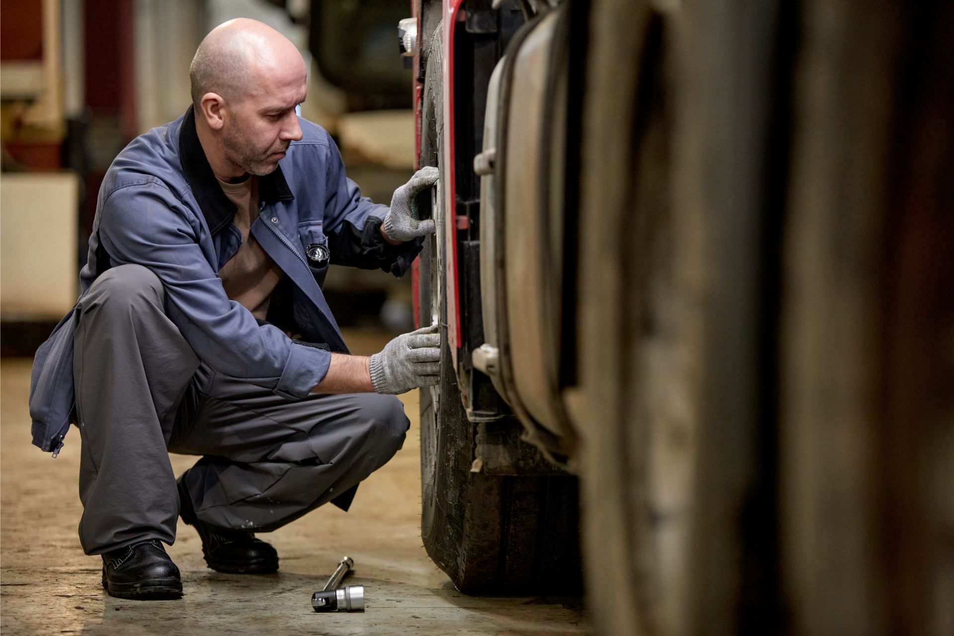 Mechanic crouching beside a car, inspecting a wheel in a garage, with tools on the floor.