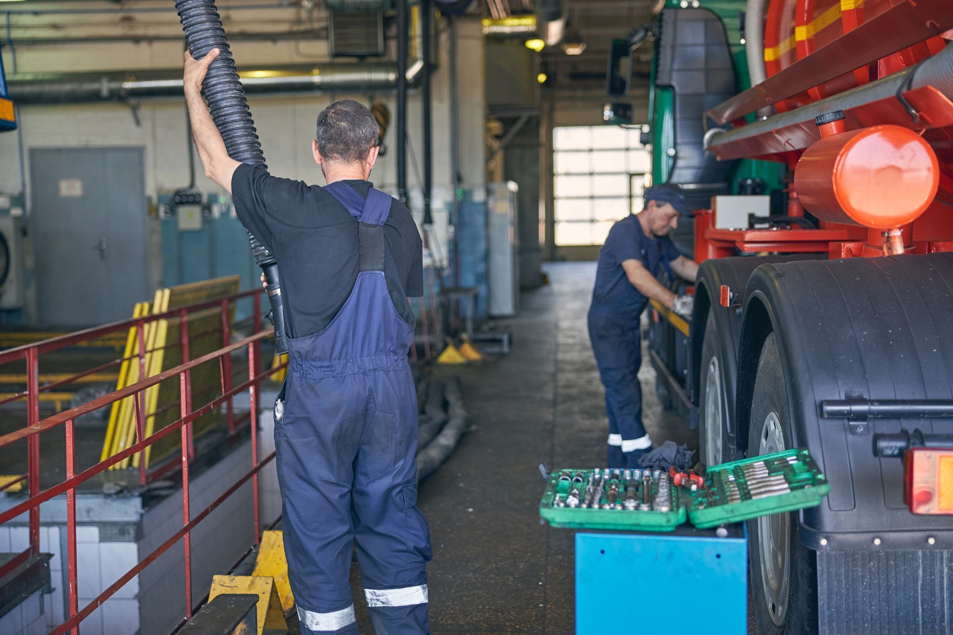 Workers servicing a truck inside an industrial garage, one standing on a platform with tools nearby