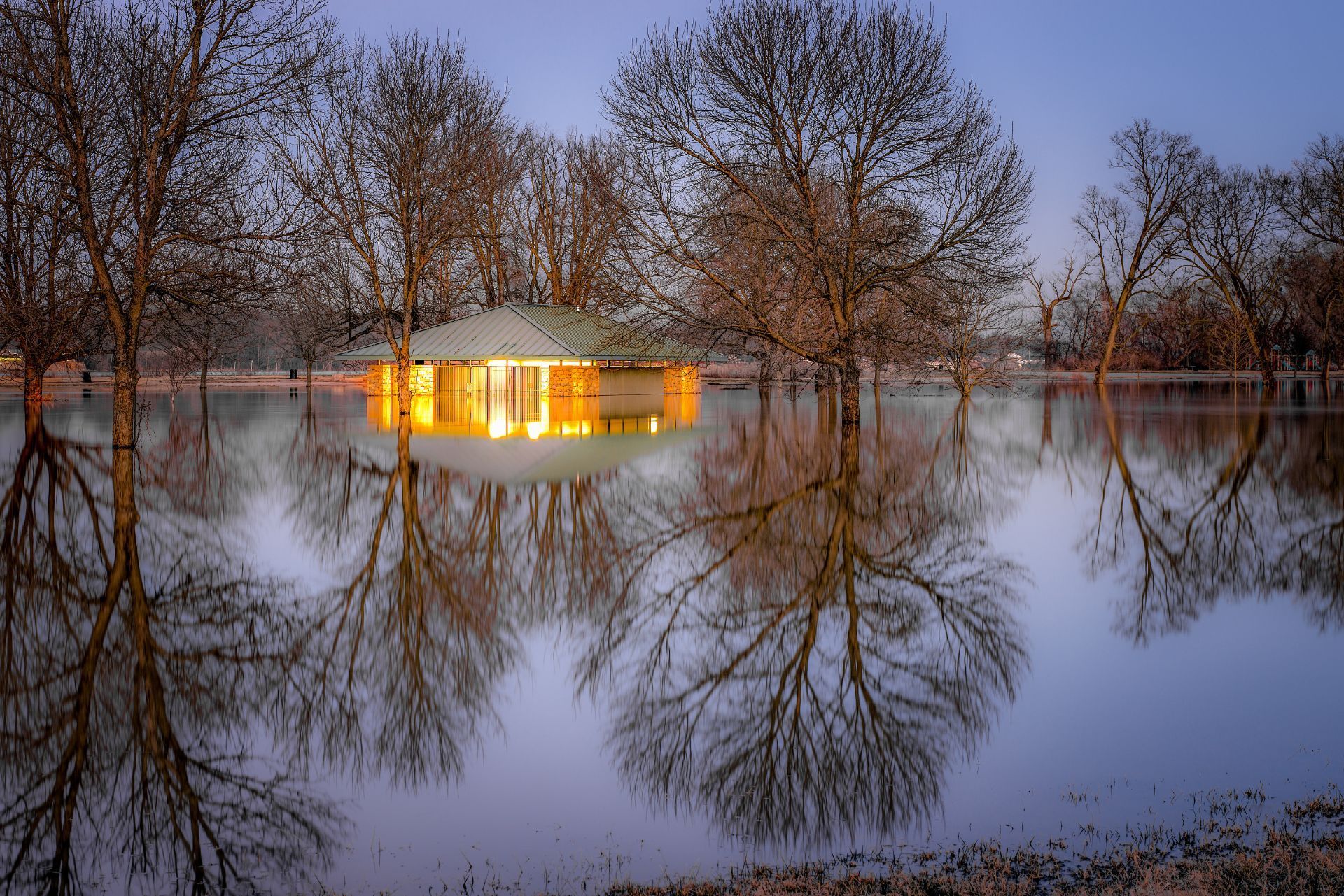 Flooded landscape at dusk, with a lit house reflected in still water and bare trees surrounding it