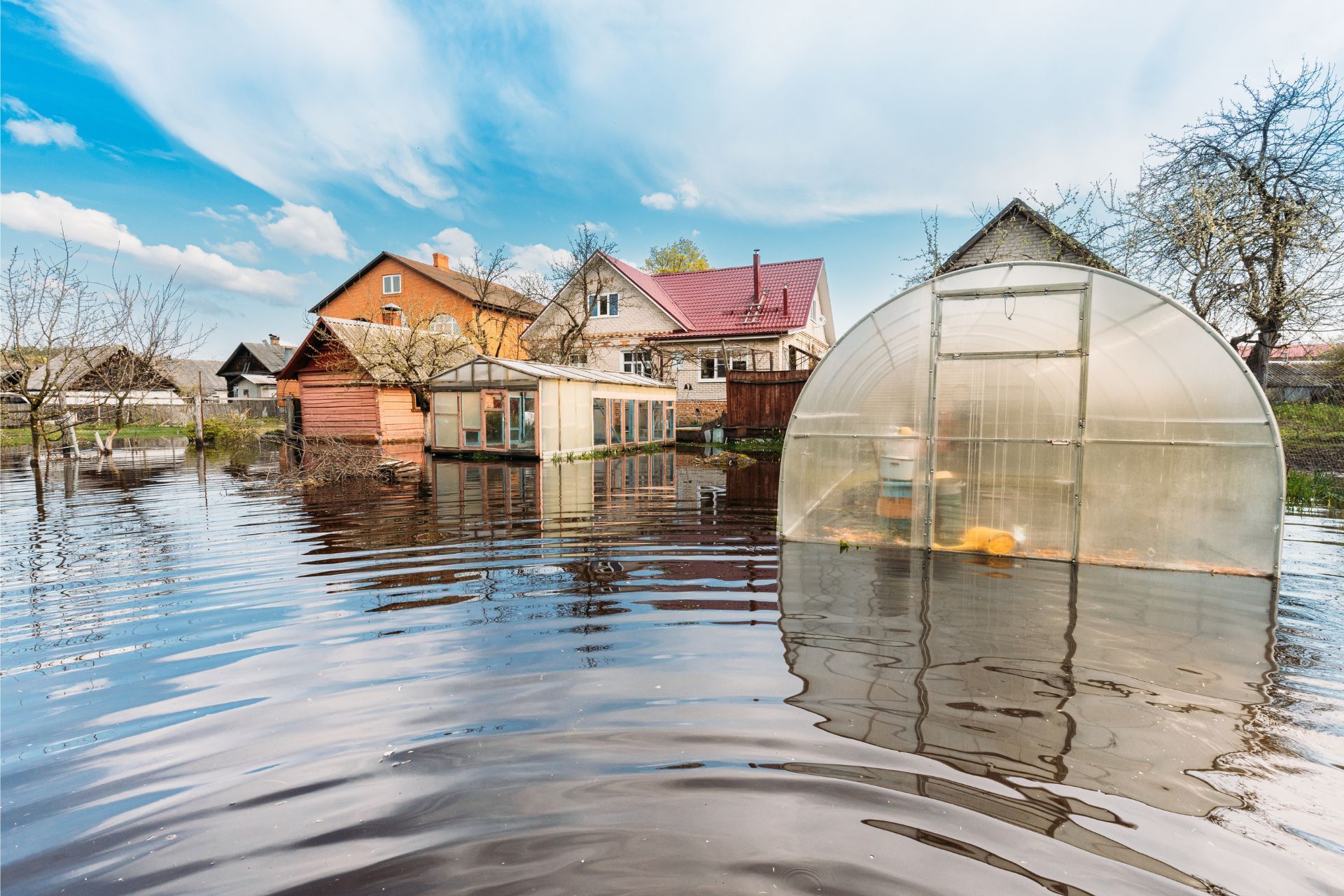 Flooded houses and a greenhouse beside a river under a partly cloudy sky