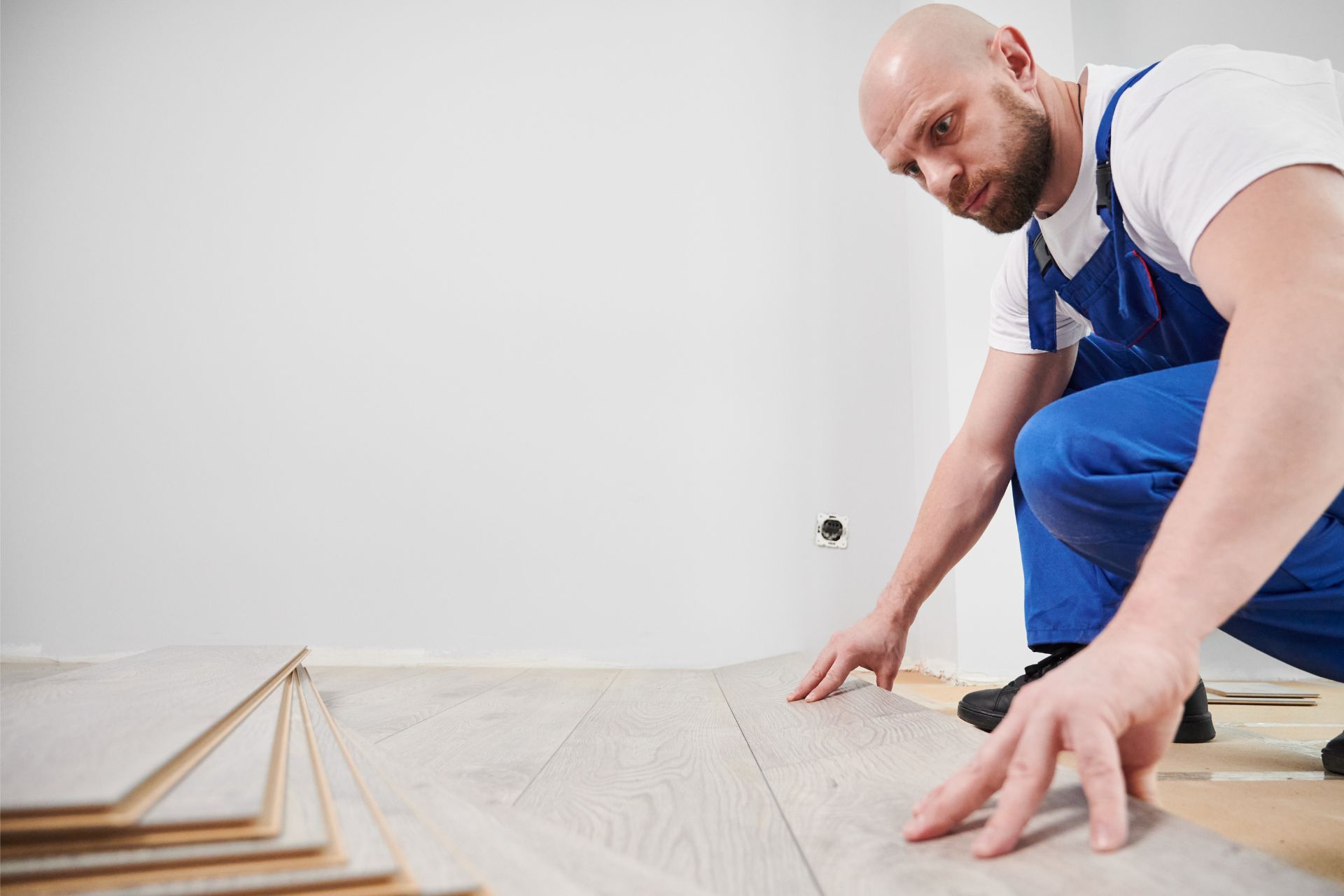 A person wearing blue work overalls kneeling and installing light-colored laminate flooring in a room with white walls.