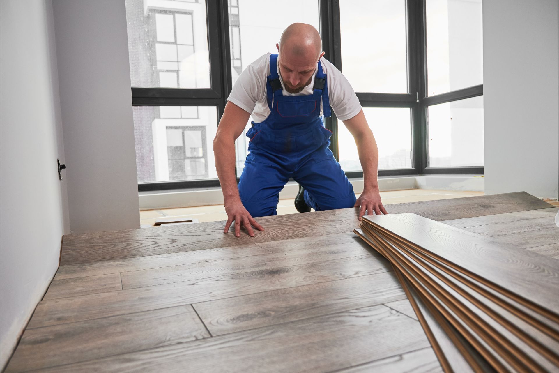 A person in blue work overalls kneels on the floor, installing light wood-look laminate flooring in a room.