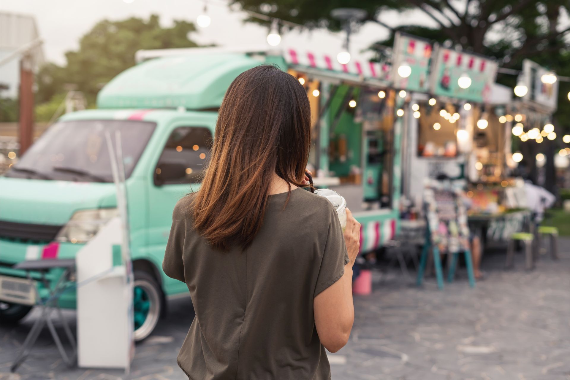 Woman seen from behind near a mint-green food truck with string lights at an outdoor market