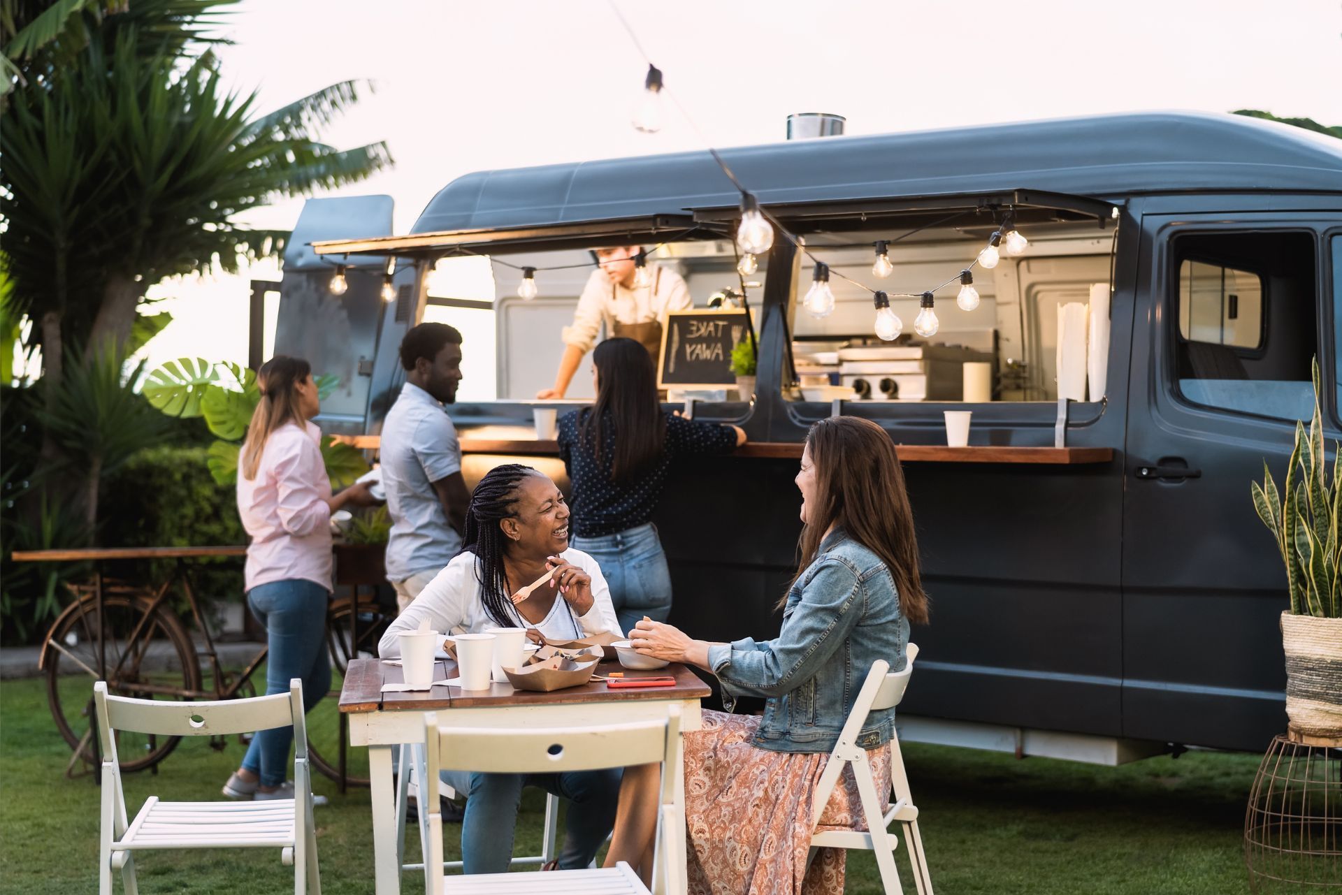 People chatting at an outdoor table beside a dark camper van at dusk.
