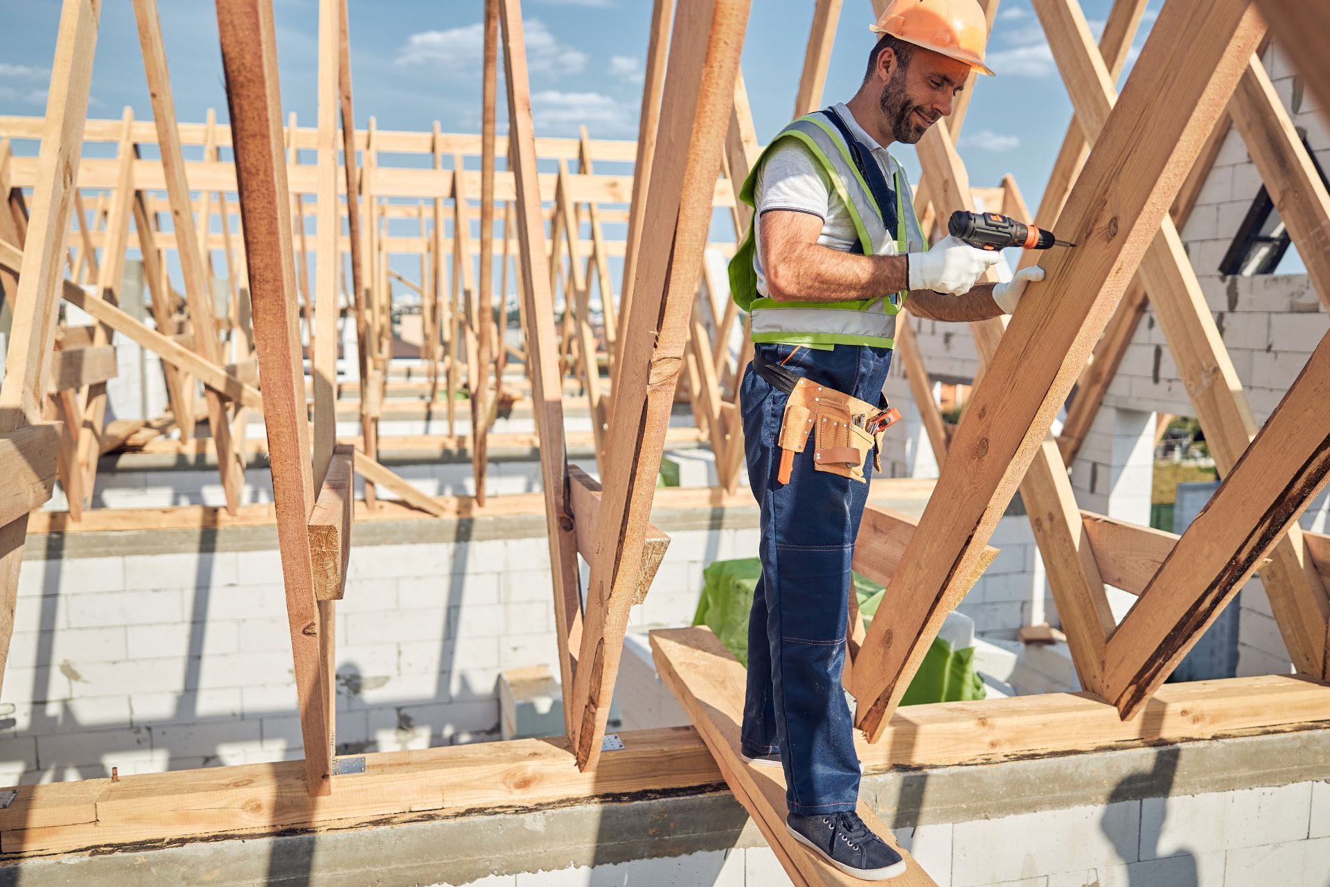 A construction worker in a hard hat and safety vest uses a drill to secure wooden roof trusses at a building site.