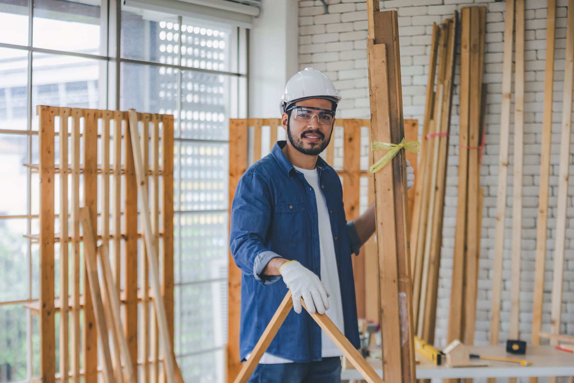 A smiling carpenter in a hard hat, goggles, and work gloves stands in a workshop, holding a large wooden board.
