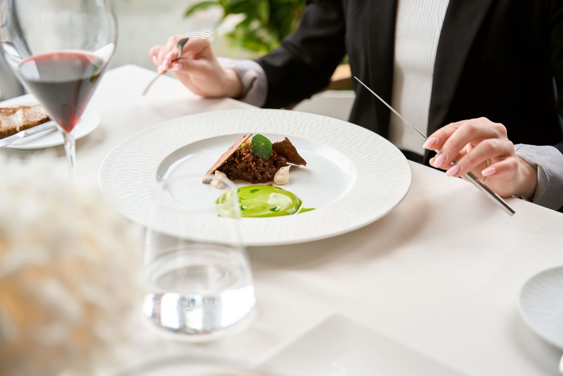A person in a black blazer dining at an upscale restaurant, preparing to eat a small gourmet dish with silver cutlery.