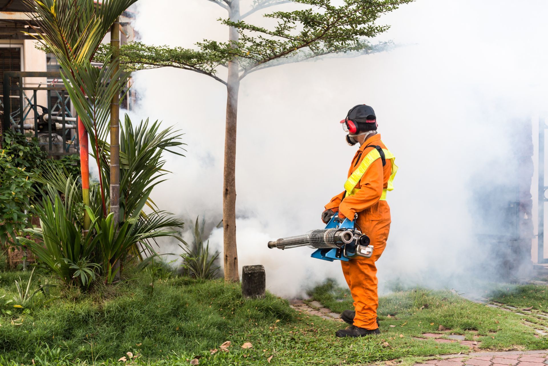 Worker in orange protective gear using a fogging machine beside a house, with thick smoke in the yard.