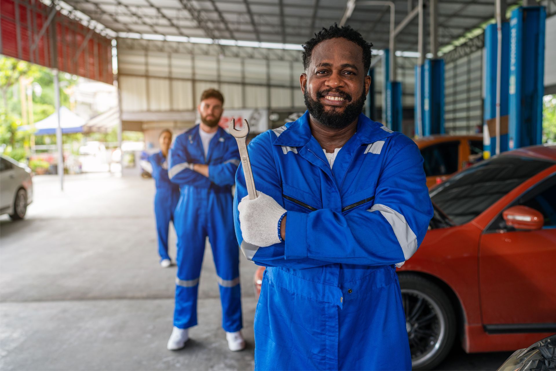 A smiling mechanic in blue coveralls and gloves stands in a workshop with two colleagues in the background.